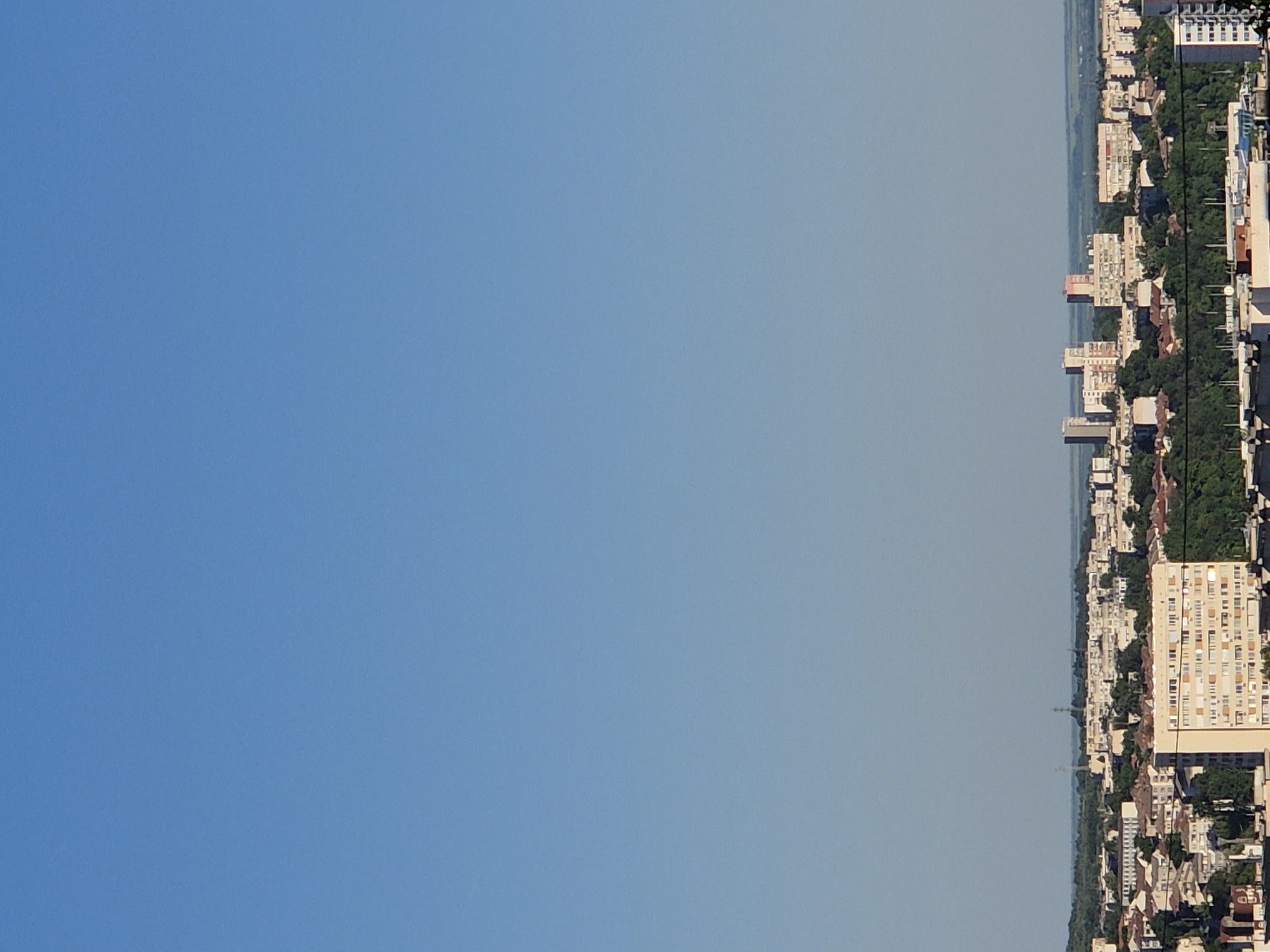 The image depicts an aerial view of an urban landscape, possibly a coastal city. Buildings of various sizes and architectural styles line the edge of a body of water, separated by dense green vegetation, likely trees. The skyline varies in height, with a mix of low-rise residential buildings and taller high-rise structures. A power line cuts through the horizon. The remainder of the image is a clear blue sky. The image is inverted.