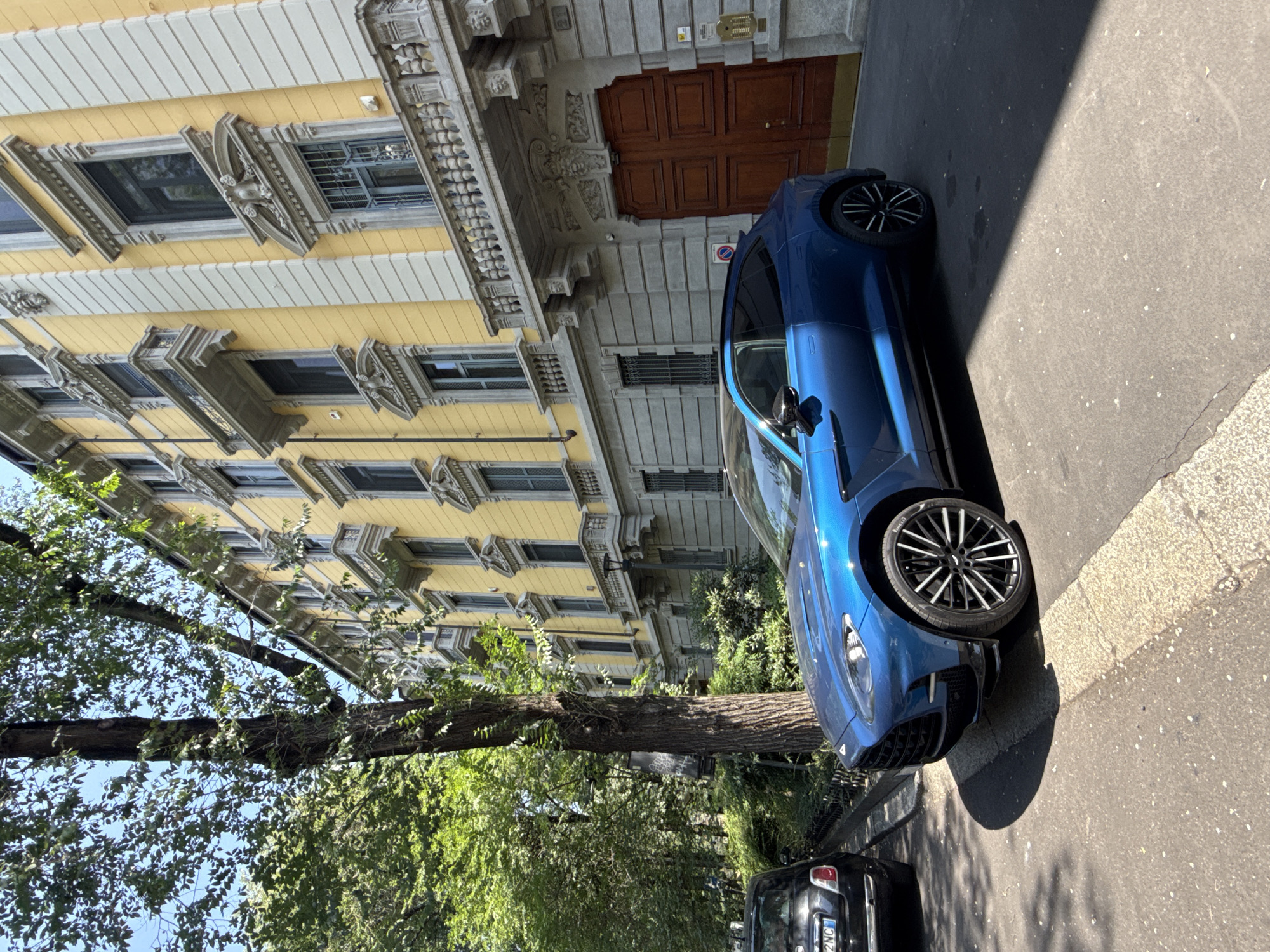 The image depicts a street scene in Milan, Italy, featuring a blue sports car parked on the side of a road. A yellow building with ornate architectural details stands behind the car. A tree with dense foliage is visible on the left side of the image. A black car is parked behind the blue car. The scene is illuminated by bright daylight.