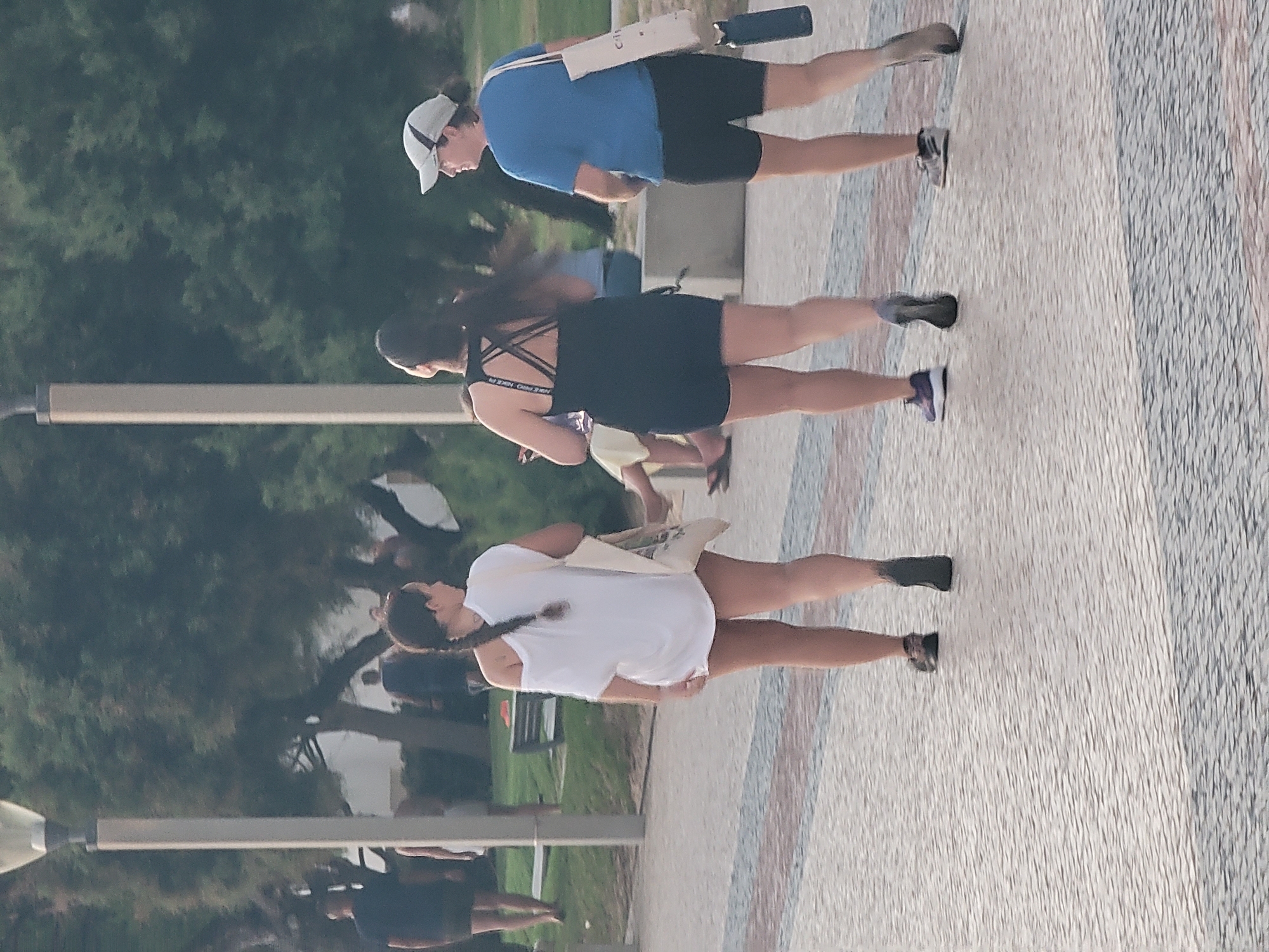 A medium shot shows three individuals walking away from the camera on a textured paved pathway in Quarteira, Portugal. The pathway is composed of small, irregular light and dark stones with subtle red and blue striped accents. To the left and background, a lamppost stands next to a lush green backdrop of trees, with some appearing to be pine trees. In the far background, hints of buildings and park benches are visible.
The person in the foreground on the left is a female with her back to the camera, wearing a loose, off-the-shoulder white t-shirt and dark shorts. Her dark hair is in a braid extending down her back. She is carrying a beige tote bag over her left shoulder.
To her right is another female, also with her back to the camera, wearing a black athletic tank top with a criss-cross back design and black shorts. Her dark hair is pulled back. She is wearing purple athletic shoes.
On the far right is a female wearing a blue t-shirt, black shorts, and a grey baseball cap. She is carrying a white tote bag over her right shoulder, from which a dark water bottle hangs. She is wearing grey and white athletic shoes.
The lighting suggests it is late afternoon or early evening, with soft shadows present. The overall atmosphere is casual and outdoorsy.