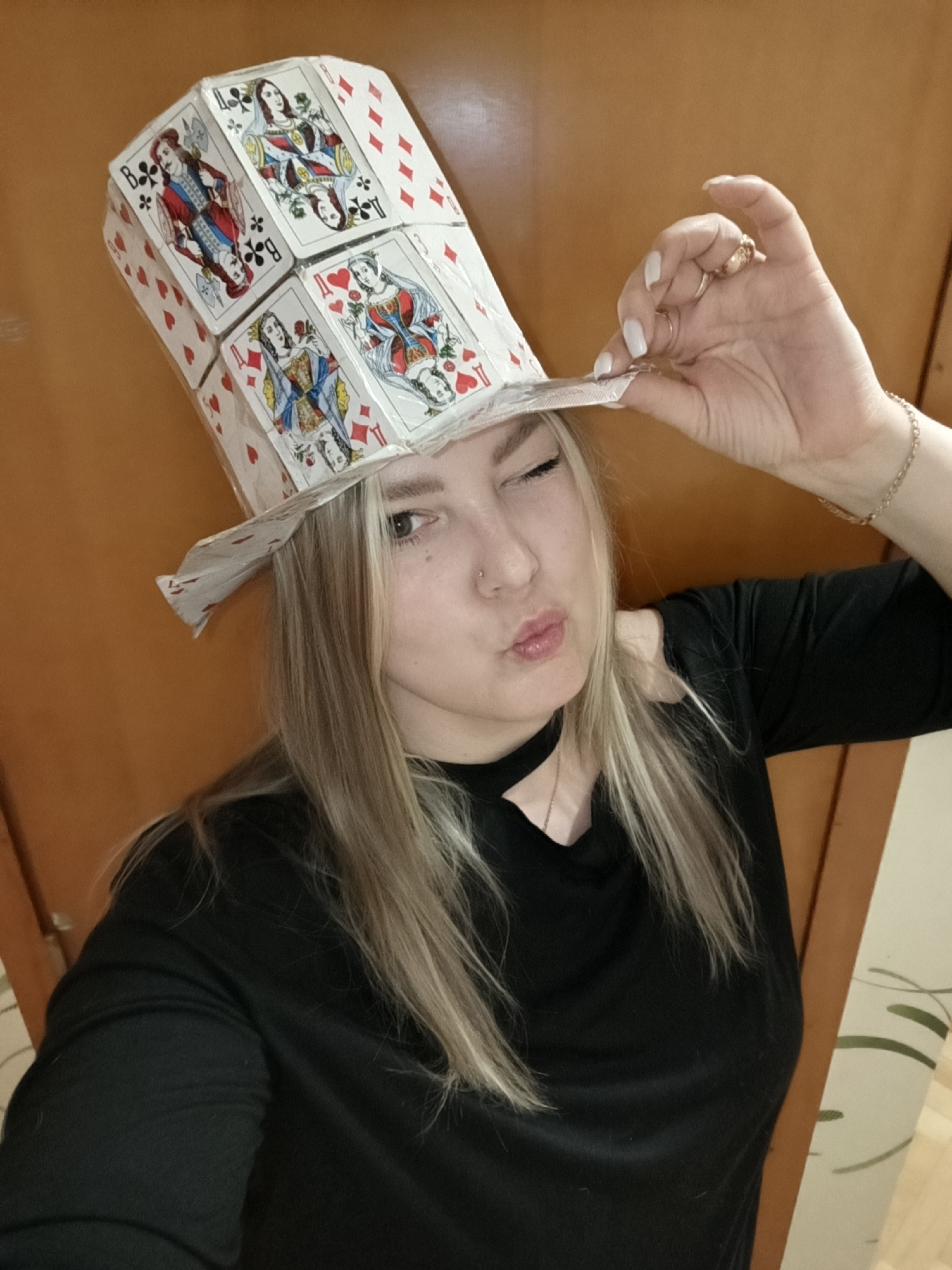 A young woman is posing indoors, wearing a top hat constructed from playing cards, in the city of Марковский, Russia. She is tilting the hat slightly with her right hand and winking, with her lips pursed. The woman has blonde hair and wears a black top with a choker necklace, a gold bracelet and rings. The background consists of a wooden door and a wall with patterned wallpaper.