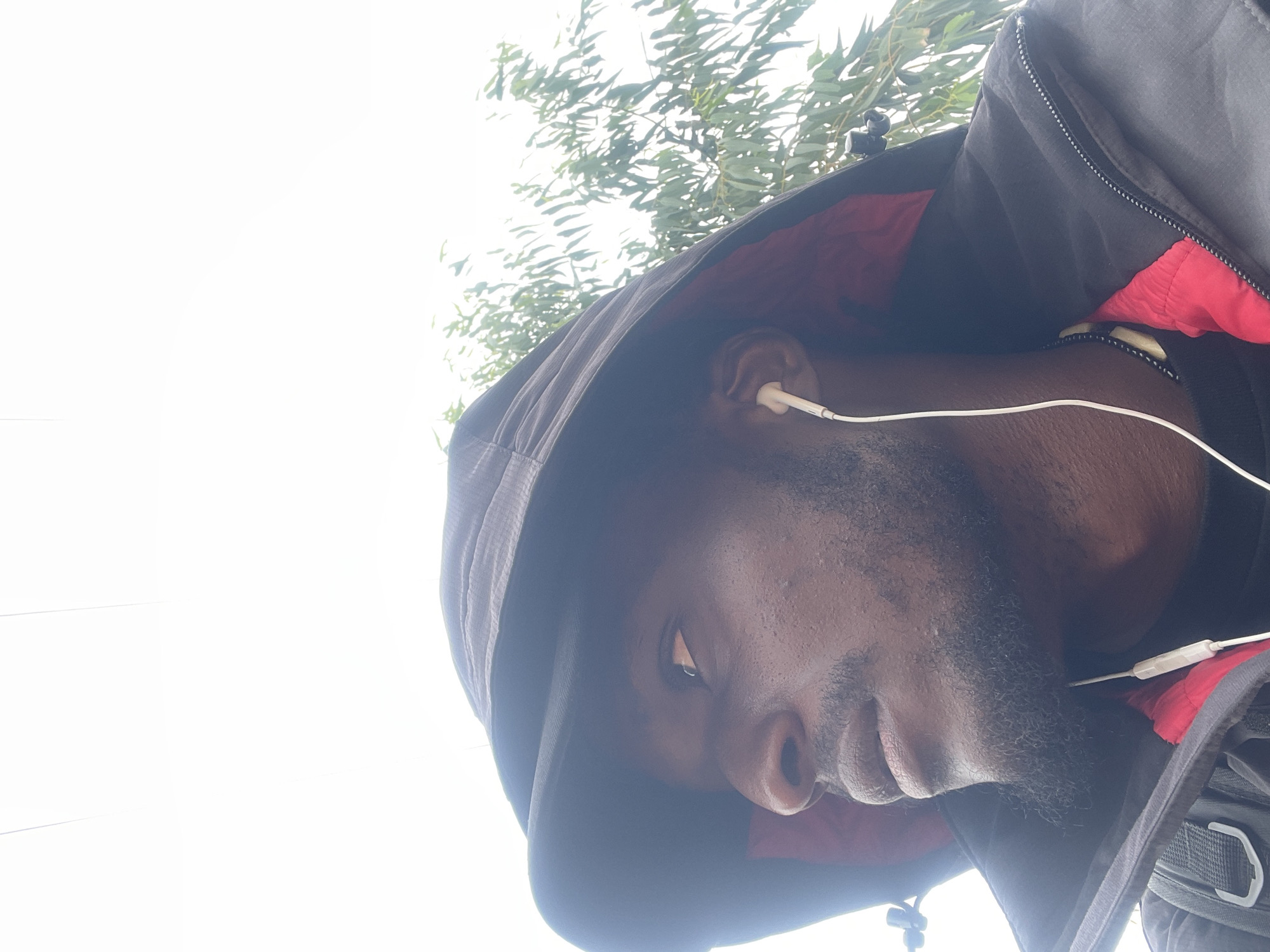 The media file is a close-up, low-angle selfie of a dark-skinned man wearing a black hooded jacket with a red lining. He has short, dark hair and a beard. He is wearing white earphones, with the wire visible across his chest. The man is looking downwards and to the left, with a neutral expression. The background consists of bright white sky and the leaves of a tree, suggesting an outdoor setting, possibly in Nigeria, given the location context. The lighting is bright, indicating it might be daytime. The image is oriented sideways.