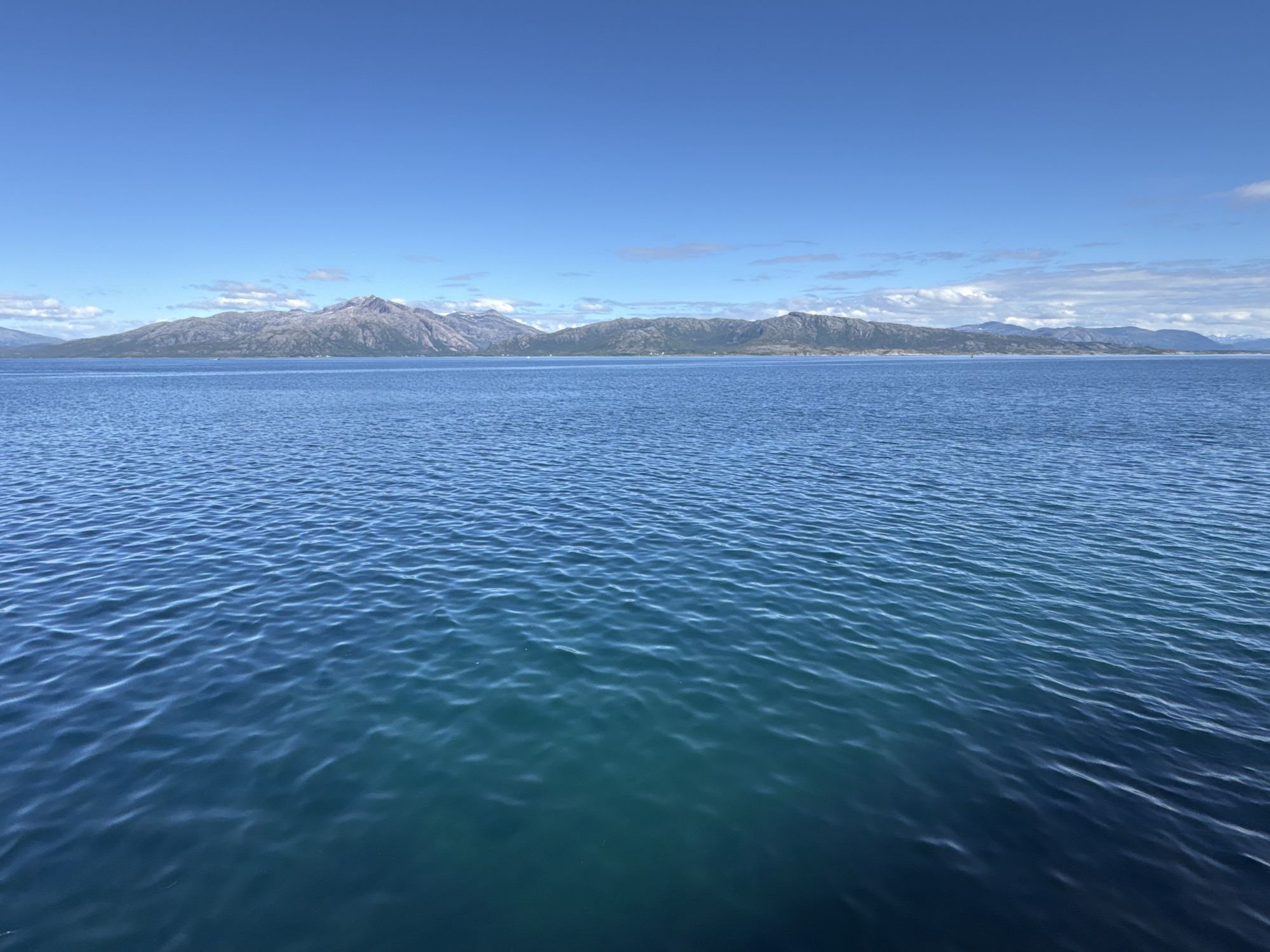 This is a landscape photograph taken from the water, looking towards a mountainous coastline under a clear blue sky. The foreground is dominated by the rippling surface of the ocean, with varying shades of blue and turquoise indicating depth. Gentle waves create a textured pattern across the water.
In the background, a long, rugged mountain range stretches across the horizon. The mountains appear barren and rocky, with some patches of snow or lighter-colored rock visible on their peaks. The coastline is irregular, with inlets and promontories. The sky is a vibrant, cloudless blue, with a few wispy white clouds scattered high up near the horizon.
The scene suggests a bright, sunny day, likely during the daytime. The clarity of the sky and the calm water indicate fair weather. The presence of mountains and a large body of water points to a coastal or fjord region. Given the provided location context, this is likely a view of the Norwegian coast, specifically Lødingen, Norway, characterized by its dramatic mountainous scenery meeting the sea. There are no visible people or specific objects that draw attention, allowing the focus to remain on the natural landscape. The overall mood is serene and majestic, emphasizing the grandeur of nature. No text is visible in the image.
