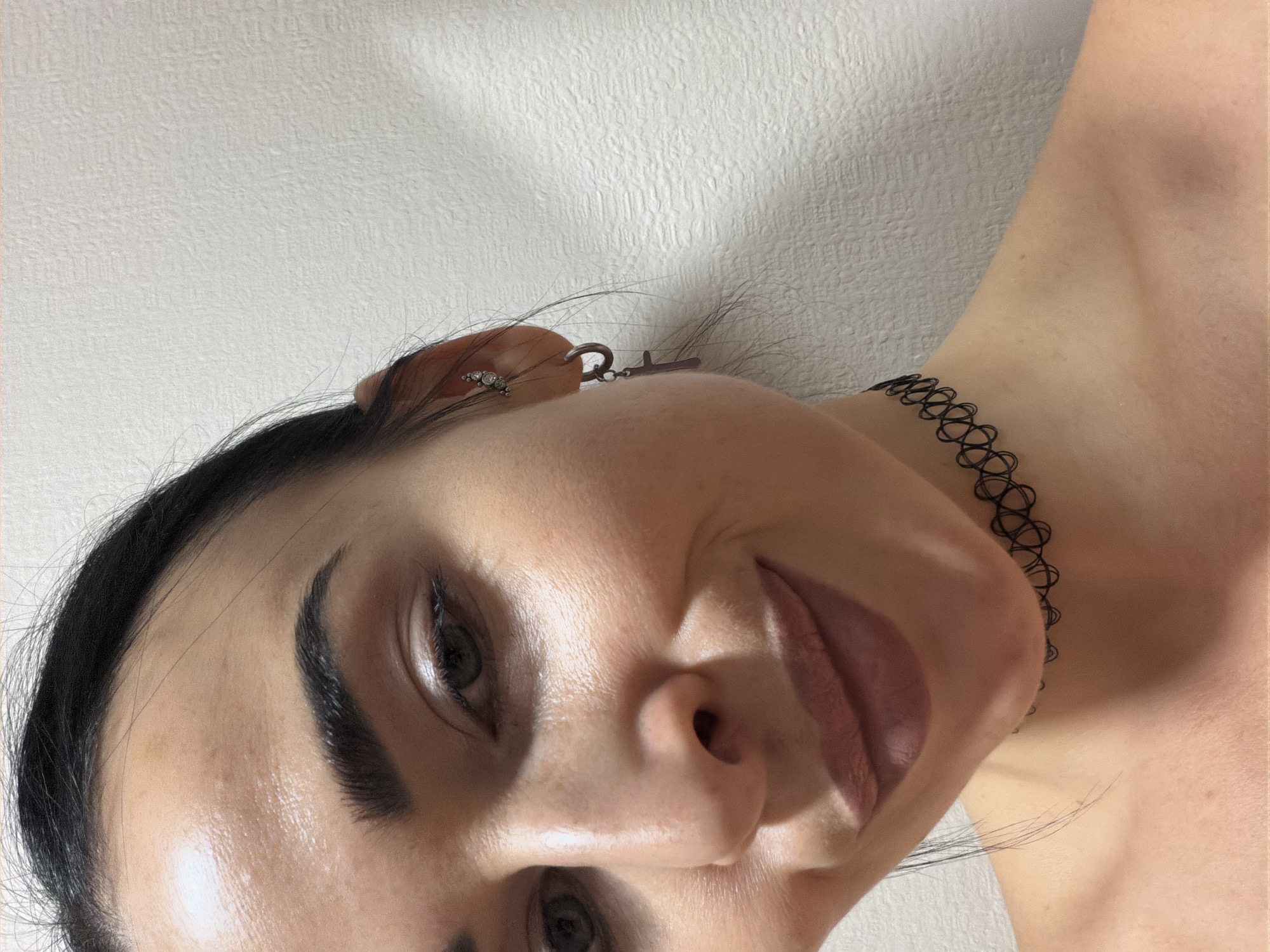 A close-up portrait shows a woman, rotated 90 degrees counter-clockwise, looking directly at the camera with a slight smile. She has dark hair, dark, well-defined eyebrows, and light skin. Her face is made up with what appears to be eyeshadow and lipstick. In her visible right ear, she wears an earring that features multiple small, clear stones and a charm resembling a key or a small cross. Around her neck is a black, intricate choker necklace. The lighting highlights her features, creating some subtle shadows, particularly under her nose and chin. The background consists of a plain, light-colored wall with a textured finish. There is no visible text in the image.
