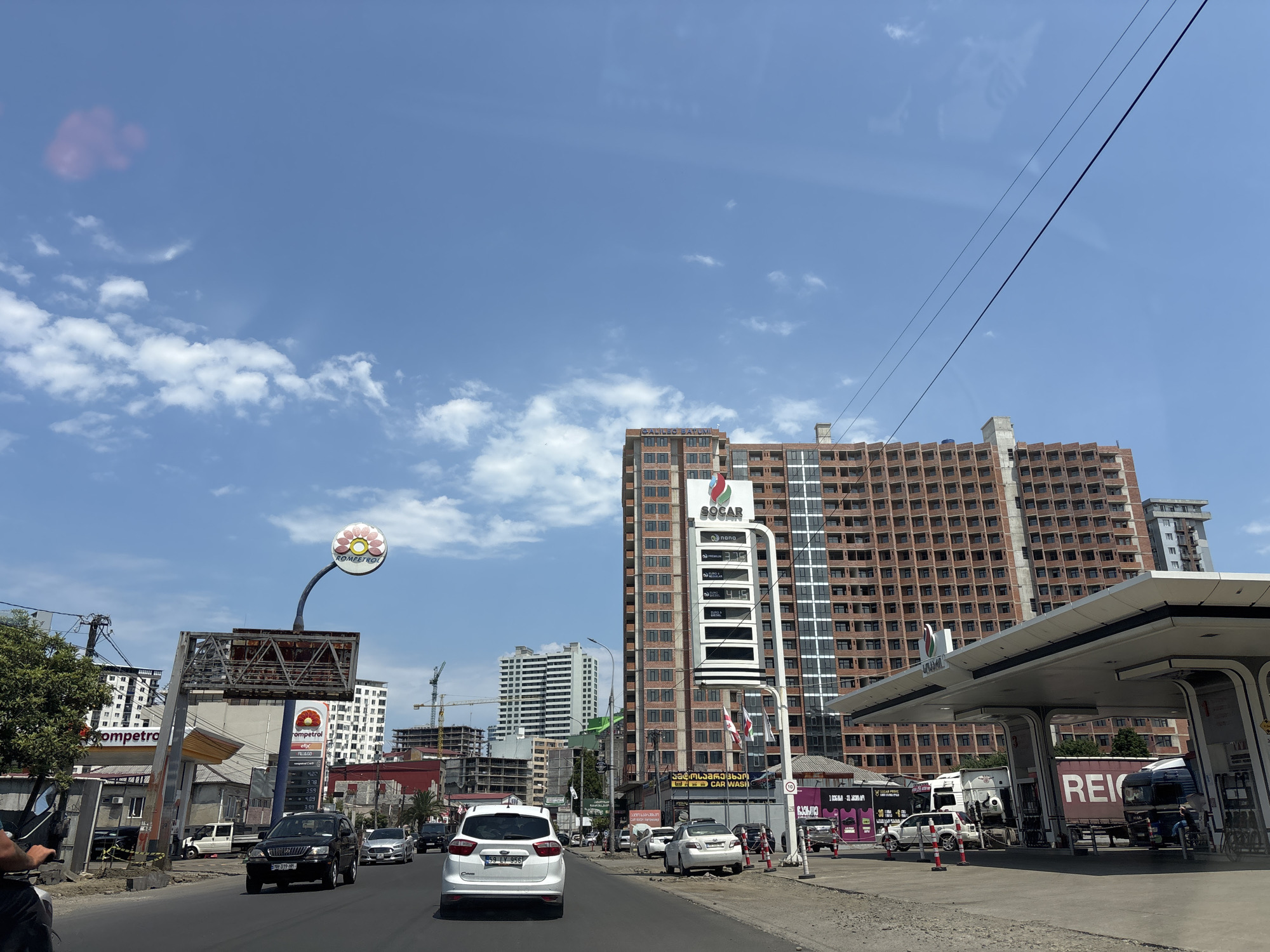 This is a street-level, point-of-view shot from a vehicle in Batumi, Georgia. The scene depicts a busy urban environment with a clear blue sky and scattered white clouds. Dominating the background is a large, multi-story brick building under construction, with many windows visible. In the foreground, a modern gas station with a white canopy is present. Several vehicles, including cars and trucks, are on the road and at the gas station. Traffic signs and streetlights are visible, along with commercial signage for businesses such as Rompetrol and SOCAR. The road is paved and appears to be a main thoroughfare, with multiple lanes of traffic. The overall impression is of a developing city with ongoing construction and active commerce.