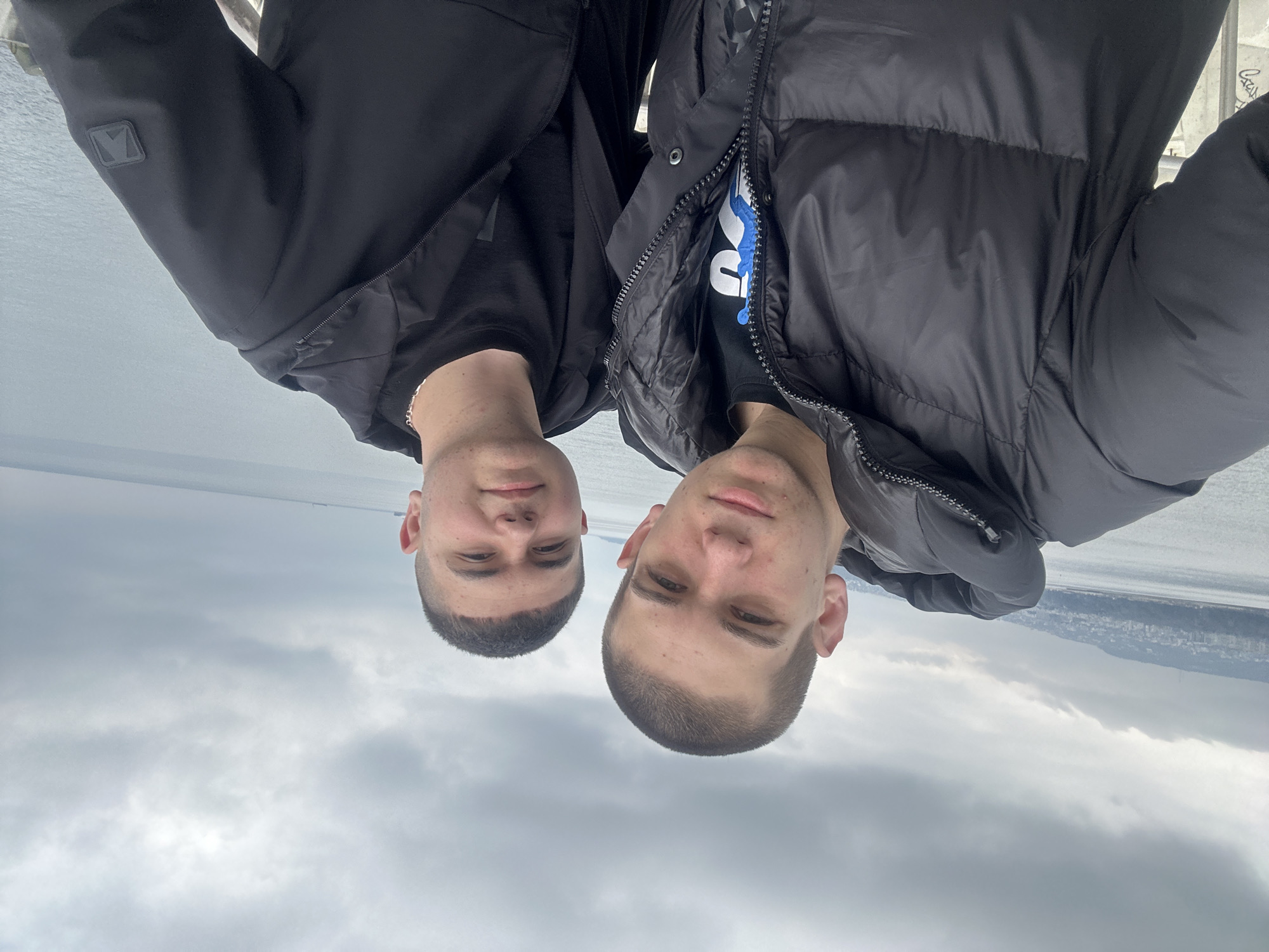 Two young men stand side-by-side outdoors, oriented upside down in the frame. They are wearing dark jackets, with one appearing to be a puffer style. The background shows a cityscape along a body of water beneath a cloudy sky. The city and location are unknown.