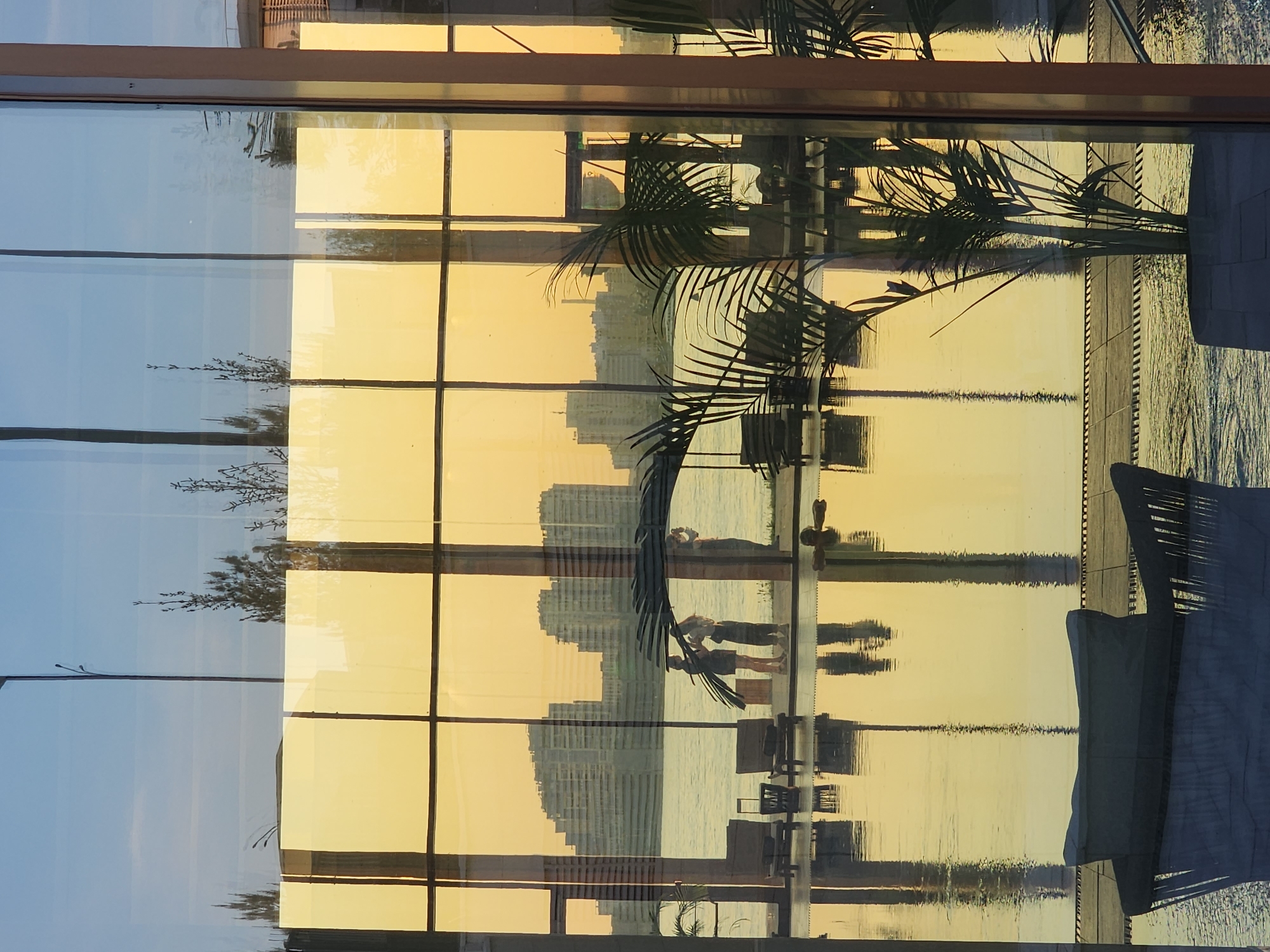Here is a detailed description of the image:
The image shows a view through a multi-paned window, reflecting an outdoor scene. The window frame is brown. The outdoor scene includes water, a cityscape in the distance, people walking along a waterfront walkway, and vegetation including palm fronds. The reflections create a layered effect, blurring the distinction between the interior and exterior. The sky appears to be a pale blue on one side and a golden yellow on the other, likely indicating sunrise or sunset. The overall setting suggests a waterfront location in a city.