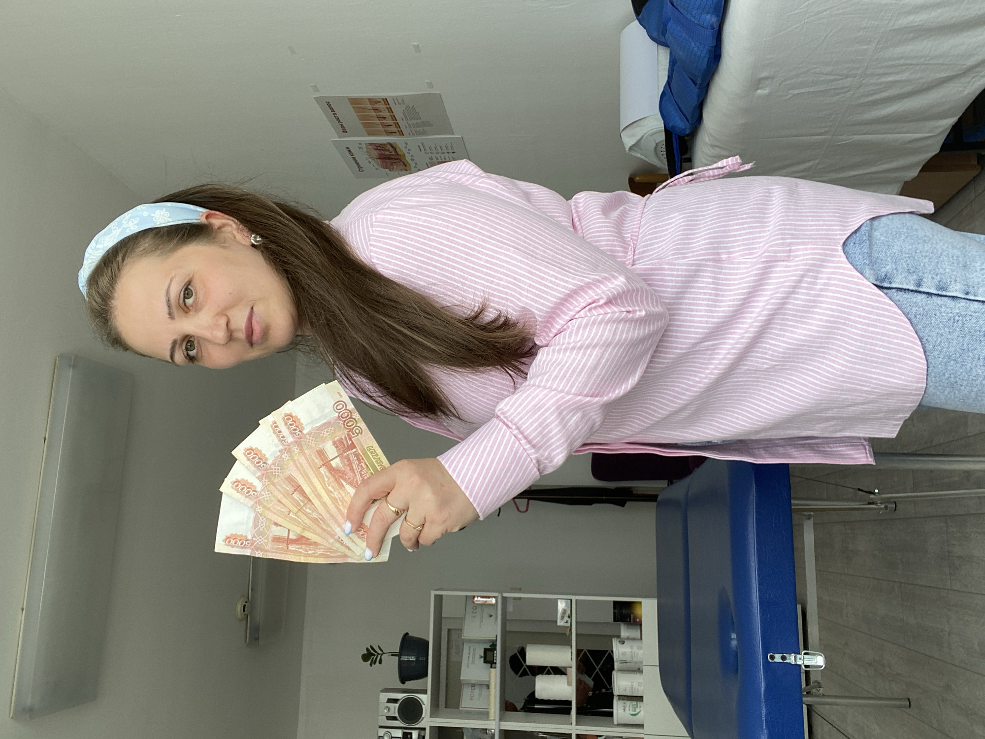 A woman is standing in a room, holding a fan of Russian 5000 ruble banknotes. She is wearing a pink and white striped button-down shirt and light blue jeans. She has a light blue patterned headband in her hair, which is pulled back into a ponytail. She is also wearing gold earrings and rings. The room appears to be a professional setting, possibly a salon or medical office, with a blue massage table in the foreground and shelves stocked with products in the background. There are also posters on the wall behind her. The location is identified as Марковский, Russia.