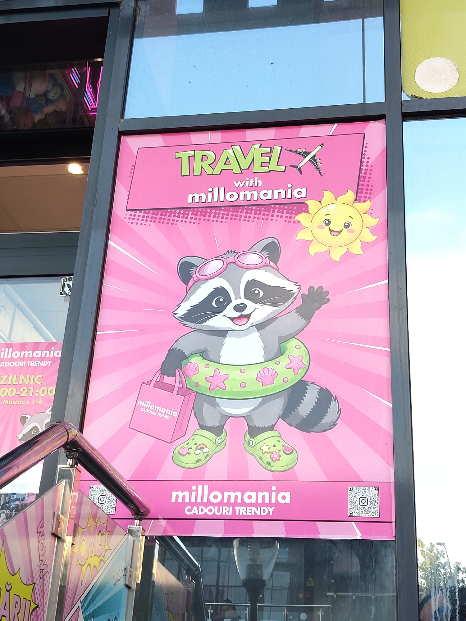 This is a close-up, eye-level, outdoor photograph of a pink advertising poster on the exterior of a building. The poster features a cartoon raccoon wearing sunglasses and a green inner tube decorated with shells and stars. The raccoon is holding a pink shopping bag with the "millomania CADOURI TRENDY" logo on it and is wearing green Crocs-style shoes. Above the raccoon, the poster reads "TRAVEL with millomania" in stylized lettering, with a cartoon sun smiling in the upper right corner and an airplane graphic on the left. At the bottom of the poster, "millomania CADOURI TRENDY" is written again, along with a QR code.
To the left of the main poster, a portion of another advertisement is visible, featuring the text "millomania," "ZILNIC 00-21:00," and "Moscova 1/4." There is also a partial image of another cartoon raccoon. Metal railings and a glass pane are in the foreground. The building appears to be a commercial establishment, potentially a shop or mall, given the advertising. The presence of a street light and trees in the background suggests an urban environment.
Based on the text "Moscova 1/4" which is a common street address format in Chisinau, Moldova, and the overall context of a commercial advertisement, this image is likely located in the city of Chișinău, Moldova.