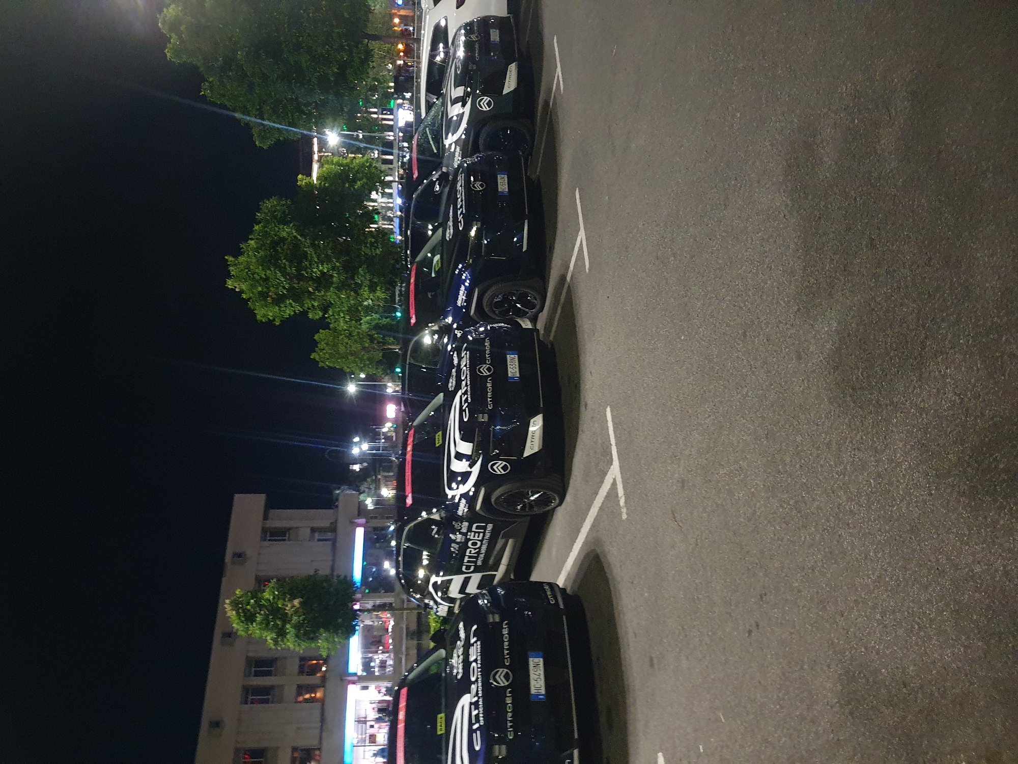 The image shows a row of black Citroën vehicles parked in a parking lot at night. The cars have white graphics and text, including the word "Citroën" and phrases like "Official Mobility Partner". Some cars have license plates visible, like "HC-549NC" and "HC-538NC".
The parking lot is marked with white lines. Behind the cars are trees, streetlights, and a building illuminated with blue and white lights. Based on the location indications in the image's metadata, it's likely the city of Plovdiv, Bulgaria. There are no visible people in the image.