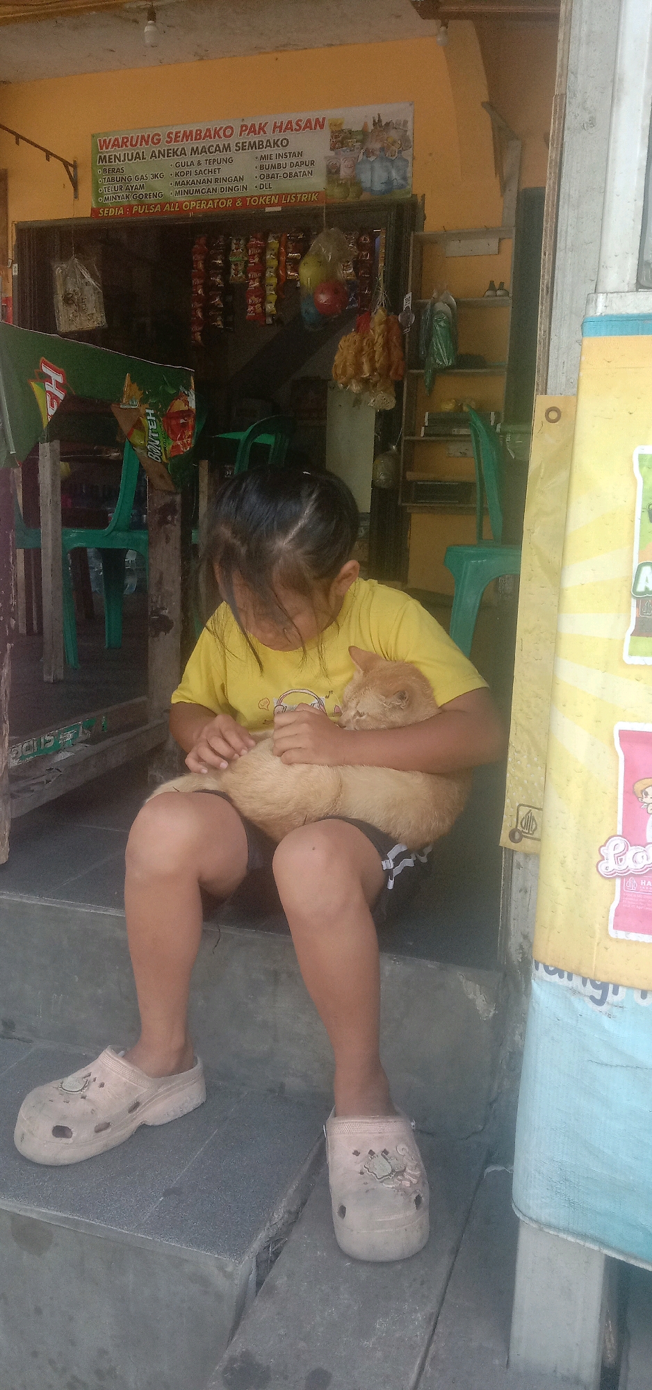 A young girl, wearing a yellow t-shirt and dark shorts, sits on concrete steps, cradling a light brown cat in her arms. The girl's hair is dark and pulled back, obscuring her face as she looks down at the cat. She is wearing light-colored slip-on shoes with holes and a design on the front. The background depicts the exterior of a small shop with a banner advertising "WARUNG SEMBAKO PAK HASAN" (Pak Hasan's Groceries) which lists various food items and services. Behind the girl, green plastic chairs are visible. To her right, a large yellow banner with some text and graphics is attached to a pillar. The overall environment appears to be a street or roadside stall in Palasari, Indonesia.