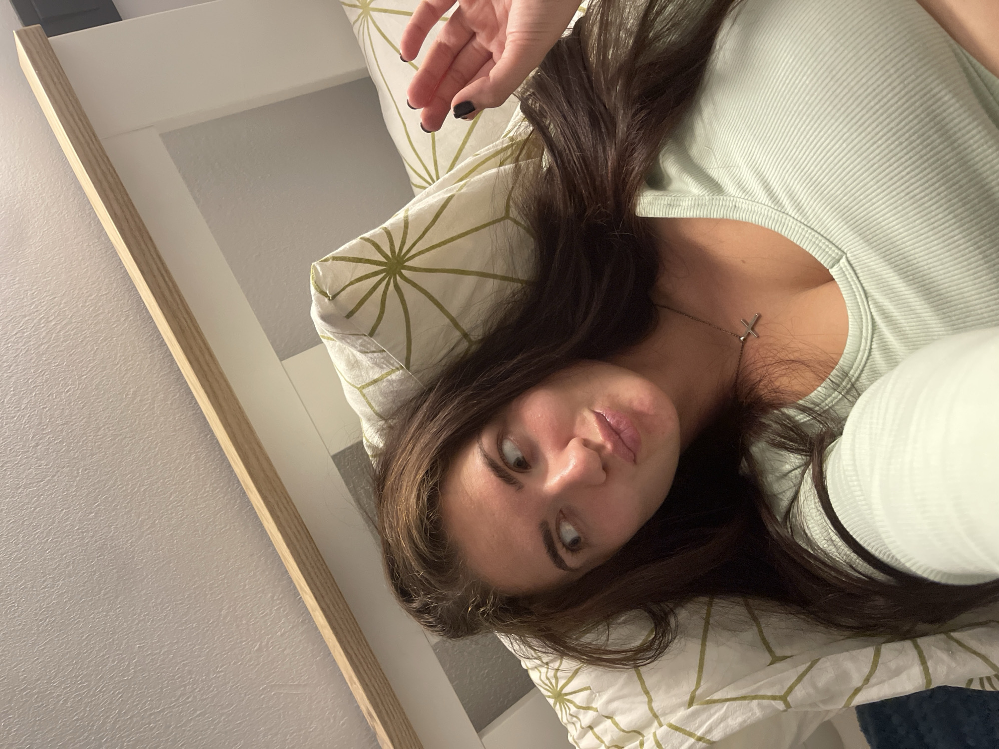 A young woman with long, dark hair is lying on a bed, propped up against pillows with a green and white geometric pattern. She is wearing a light green, ribbed shirt and a silver cross necklace. She is looking away from the camera with her lips pursed. A hand with black nail polish is raised near her head. The background shows a wooden headboard and a textured wall. The image appears to be taken indoors in a bedroom. There is no identifiable information or context that would place this image in Мосметаллоконструкция Онт, Russia.