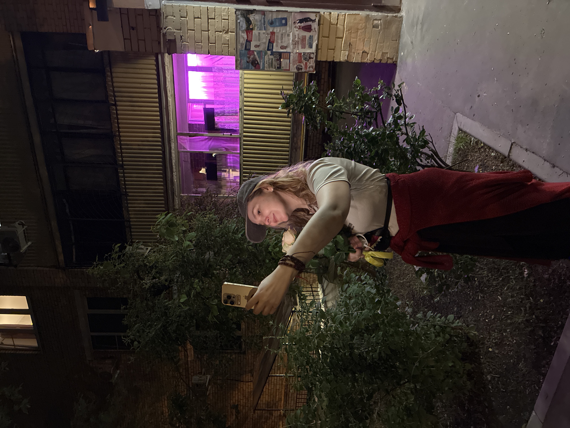A woman stands outdoors in what appears to be an alleyway at night. She is holding a gold-colored smartphone and appears to be taking a photo. She is wearing a gray baseball cap, a light-colored t-shirt, and a red shawl or wrap. Behind her are buildings with windows, some lit up with a purple glow. There is also greenery in the form of shrubs and trees present in the scene. The environment seems urban and suggests an area with both residential and commercial properties. It is not possible to confirm the exact location as Moscow, Russia, from visual data alone.