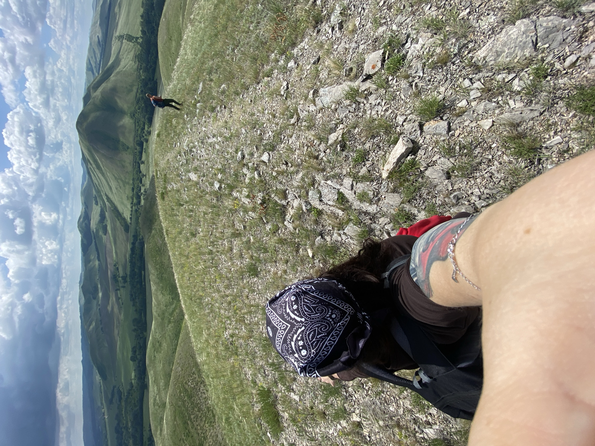 In Yuldybaevo, Russia, two individuals are hiking through a mountainous landscape under a cloudy sky. In the foreground, a person wearing a black bandana with a white paisley pattern and a brown shirt is seated, with a black backpack and a red garment visible. A tattoo is present on the person's left arm, adorned with a silver bracelet. In the mid-ground, another person with a backpack walks along a rocky path. The surrounding environment consists of rolling green hills covered in grass and scattered rocks, with trees dotting the landscape in the distance.