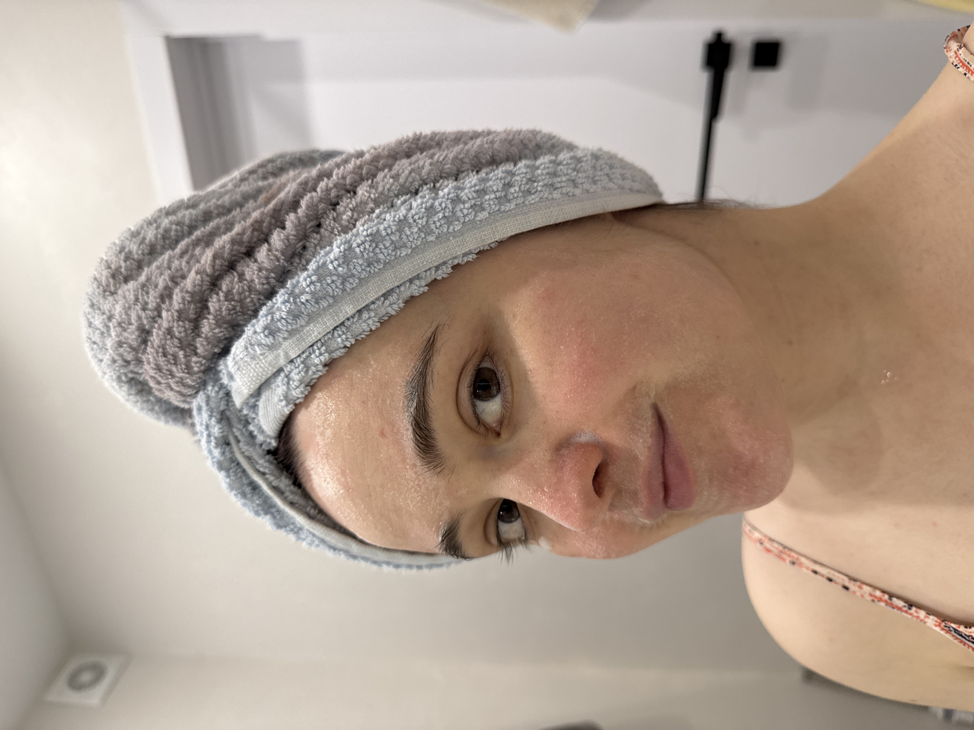 A close-up shot depicts a woman with a towel wrapped around her head, likely after showering or washing her face. The woman's face shows evidence of a skincare routine, with a light residue of a cleansing product or moisturizer visible on her skin. Her gaze is directed towards the viewer. The background is a simple bathroom setting, with white walls and a glimpse of a towel bar and a portion of a shower fixture. The shot does not feature any indications of the location being Izhevsk, Russia.