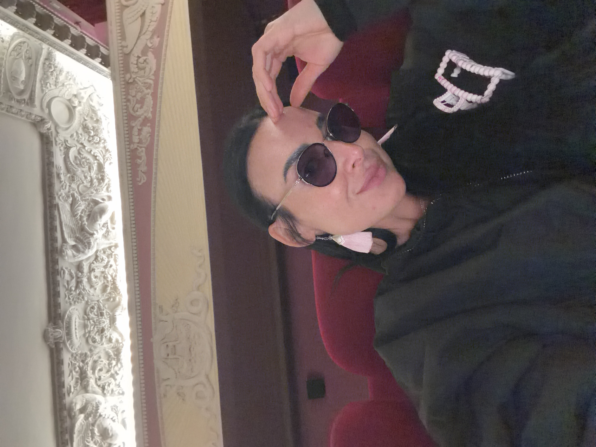 A person is captured in a medium close-up, lying on their back or reclining in a plush red seat. They have dark hair and are wearing dark, round sunglasses and a dark hooded sweatshirt with prominent white drawstrings. One hand is raised, fingers lightly touching their forehead. A light pink tassel earring is visible dangling from their ear. The person has a slight smile on their face, suggesting a relaxed and content mood.
The setting is an opulent interior, likely a historic theater, opera house, or grand hall, consistent with architecture found in Saint Petersburg, Russia. To the left, elaborate white architectural moldings and carvings adorn a wall or column. These intricate decorations feature classical elements, including what appear to be busts, floral patterns, and possibly mythical creatures like griffins or cherubs. A vertical band with pink detailing is also visible alongside the white ornamentation. The overall aesthetic suggests a lavish and ornate environment. No discernible text is visible in the image. The lighting appears to be indoor artificial light.