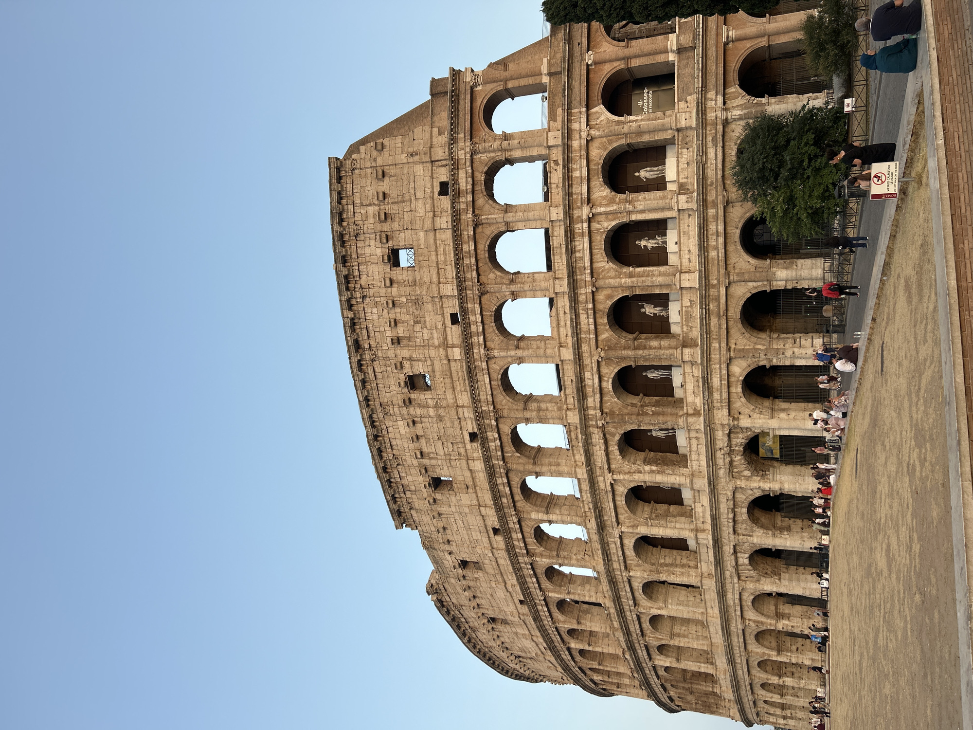 The image shows the Colosseum in Rome, Italy. The iconic amphitheater, constructed of travertine limestone and volcanic rock, is seen from a low angle, emphasizing its massive scale. The sunlit stone structure features multiple tiers of arched openings, with statues visible within some of the upper arches. A clear blue sky serves as the backdrop. In the foreground, a dry, sandy ground slopes upwards towards the Colosseum. A crowd of tourists, dressed in casual summer clothing, are gathered on the open area and along a paved pathway in front of the monument. Some individuals are seated, while others are standing and observing the structure. There are several trees and greenery visible to the right of the Colosseum, along with some fencing and signage, including a sign that appears to be a "Do Not Walk on Grass" warning. The overall scene depicts a typical daytime view of this historic landmark, bustling with visitors.