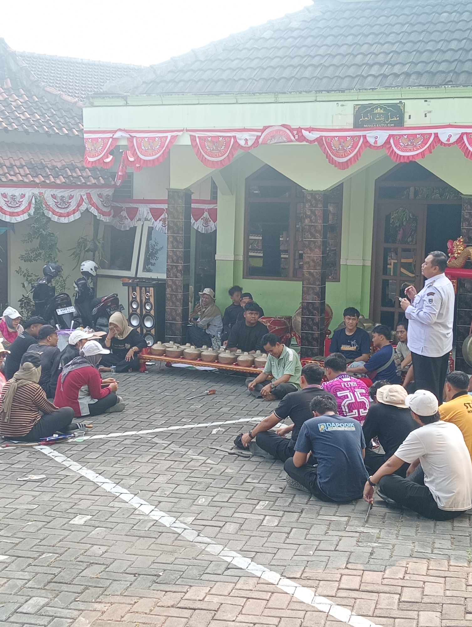 The image depicts an outdoor gathering in what appears to be a residential or community area in Demak, Indonesia. A group of people are seated on the ground in front of a building decorated with red and white festive banners. In the foreground, several individuals are seated, facing a row of musical instruments, likely a gamelan set, arranged on a wooden platform. Some individuals are actively engaged, either watching the performance or participating in the activity.
In the middle ground, a man in a white shirt and dark pants stands and appears to be addressing the seated group. Behind him, the building has a green facade with decorative pillars and windows. A sign above an entrance reads "MUSALA FAITUL ILMI" in Arabic script and English, indicating the building is a prayer hall. Several motorcycles are parked to the left of the building, suggesting this is a common mode of transportation in the area.
The ground is paved with interlocking bricks, and a white line is painted across it. The overall atmosphere suggests a cultural event or community gathering taking place during daylight hours. The weather appears overcast.