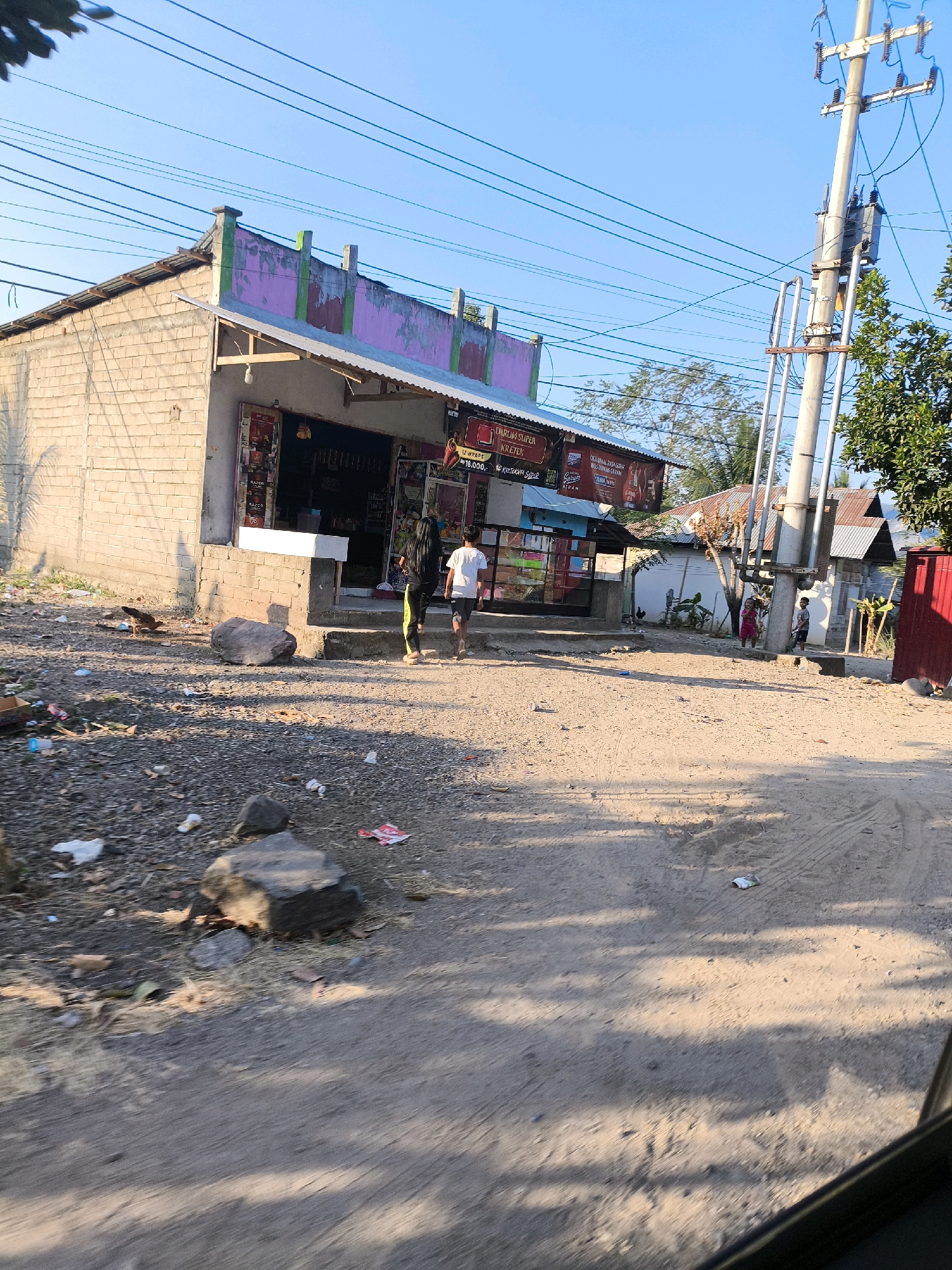 The image depicts a street scene in Dulupi, Indonesia, on a clear day. In the foreground, a dirt road is visible, with tire tracks and scattered debris, including rocks and plastic waste. A small chicken is pecking at the ground near a brick wall.
To the left of the road, a small, light-colored brick building serves as a storefront, with a pink and green painted facade. Advertising posters are displayed above the open entrance. Two individuals, a woman with long dark hair and a man in a white t-shirt, are walking away from the building towards the right.
Dominating the right side of the image is a tall utility pole with multiple electrical wires crisscrossing the sky. Further back, a cluster of buildings with corrugated metal roofs and various trees are present. A red-sided structure is also visible on the far right. The overall impression is one of a rural or semi-urban area with a simple, functional streetscape.