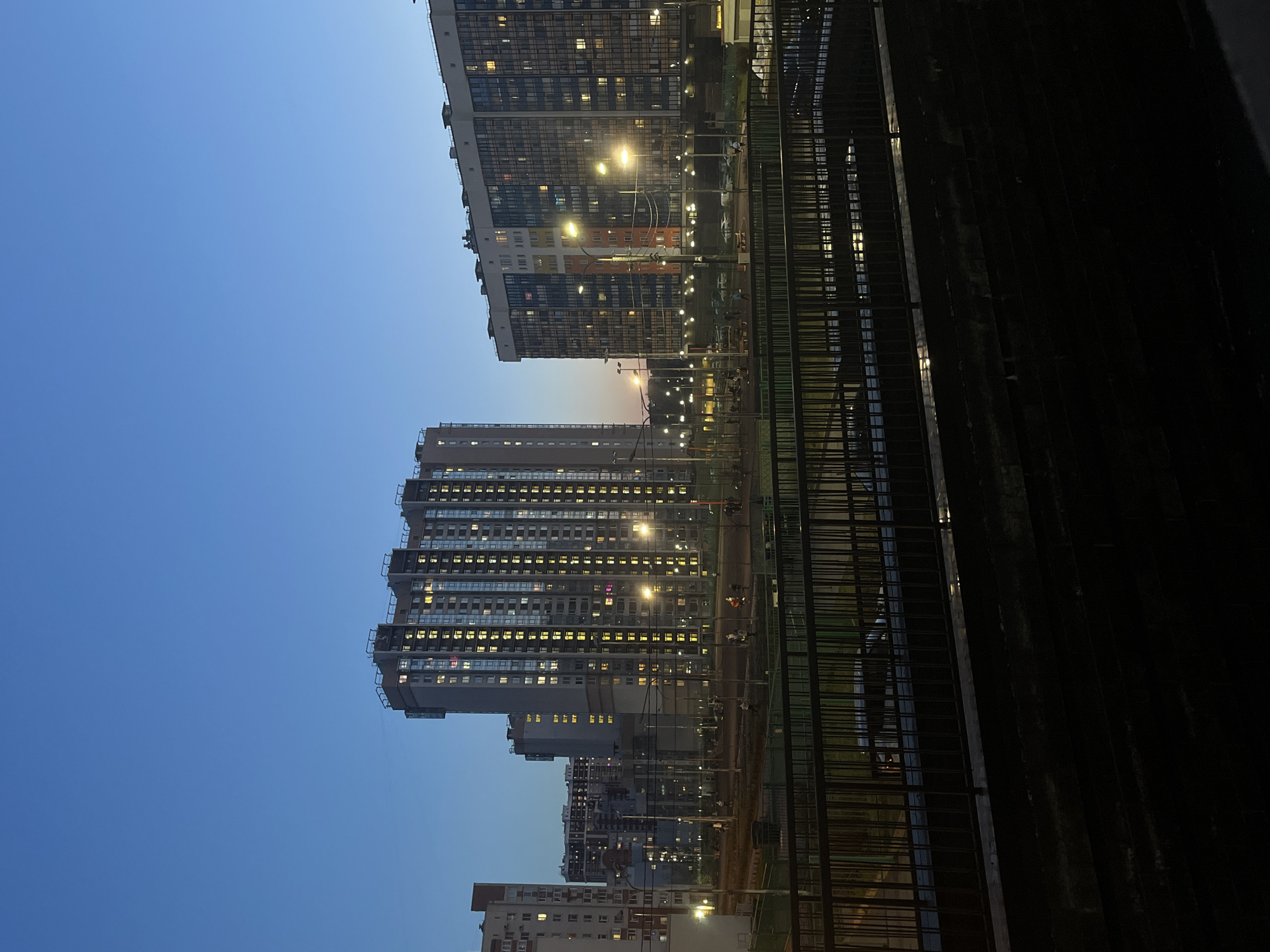 The image shows a view of apartment buildings in the city of Vsevolozhsk, Russia at dusk. The sky is a gradient of blue. The buildings are modern, high-rise apartment blocks with many lit windows. A metal railing obscures a significant portion of the foreground. The scene is illuminated by streetlights and lights from the apartment windows. The time appears to be late evening or early night.