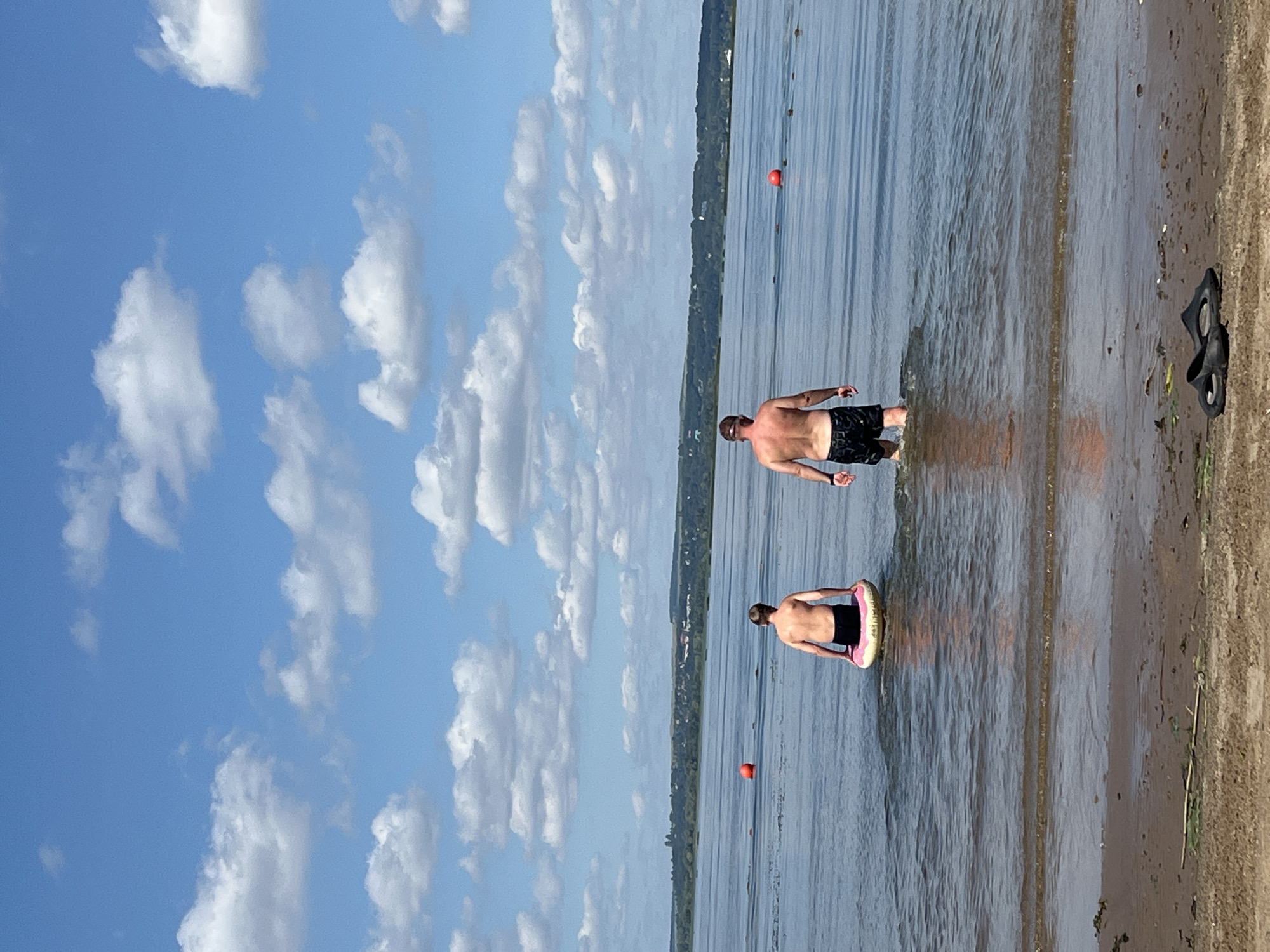 The image depicts a body of water with a sandy shore in the foreground, under a bright blue sky with scattered white clouds. Two individuals are partially submerged in the water. One person, further from the shore, is seated on a pink and yellow inflatable ring. The other person, closer to the shore, is standing in the water, facing away from the camera. In the distance, a tree-covered shoreline with some buildings is visible. Two red buoys are present in the water, and a pair of black sandals are on the sand in the foreground. The overall scene suggests a recreational setting on a warm day. The location is identified as Shumeika, Russia.