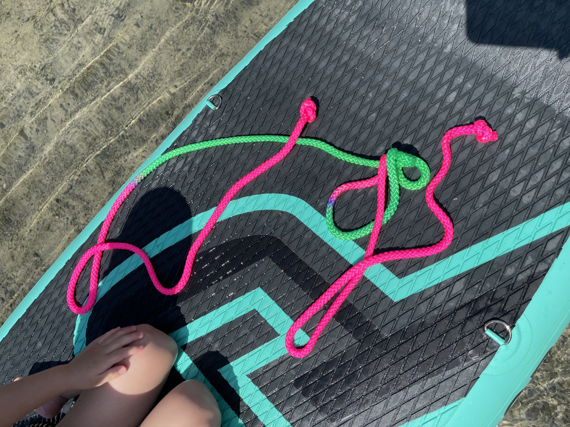 An overhead shot shows a person's legs and a paddleboard on the water. The paddleboard is black with teal accents and has a textured surface. Two brightly colored ropes, one neon pink and one neon green, are coiled on the paddleboard. The water is clear and shallow, allowing the sandy bottom to be visible. The scene appears to be a sunny day, possibly at a lake or river. There is no indication of the location being in Korosten, Ukraine.