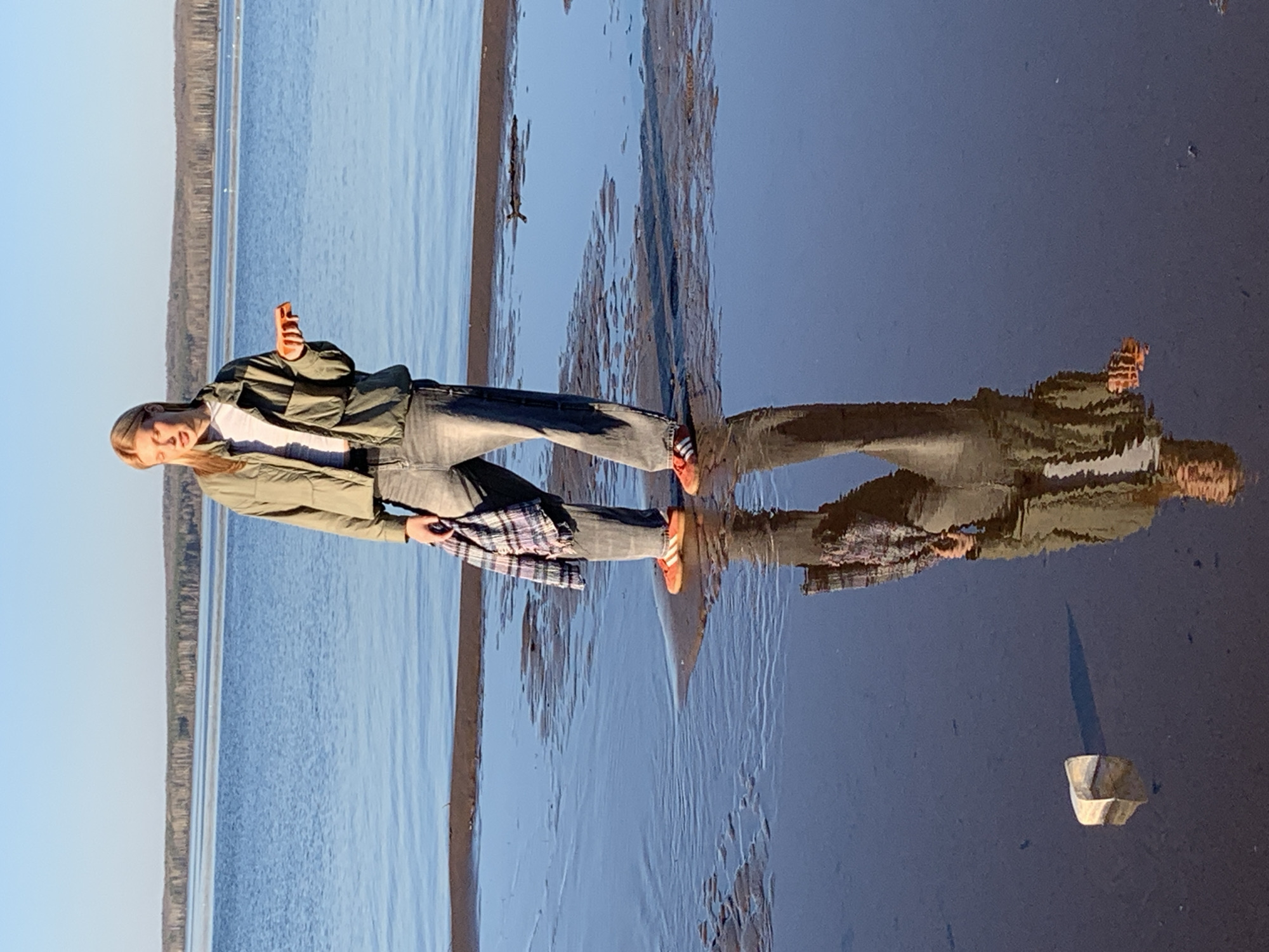 A woman stands on a beach at what appears to be a body of water. She is wearing a green jacket, grey jeans, and red and white sneakers. She is holding a cell phone in her right hand and a plaid shirt in her left hand. The water reflects her image. A small stick or piece of debris is floating in the water near the woman. In the distance, there is a cliff or elevated land feature. Debris is on the ground next to the water. The location is likely Chaikovsky, Russia, based on the city requested. The image is taken in landscape mode but appears to be rotated 90 degrees clockwise.