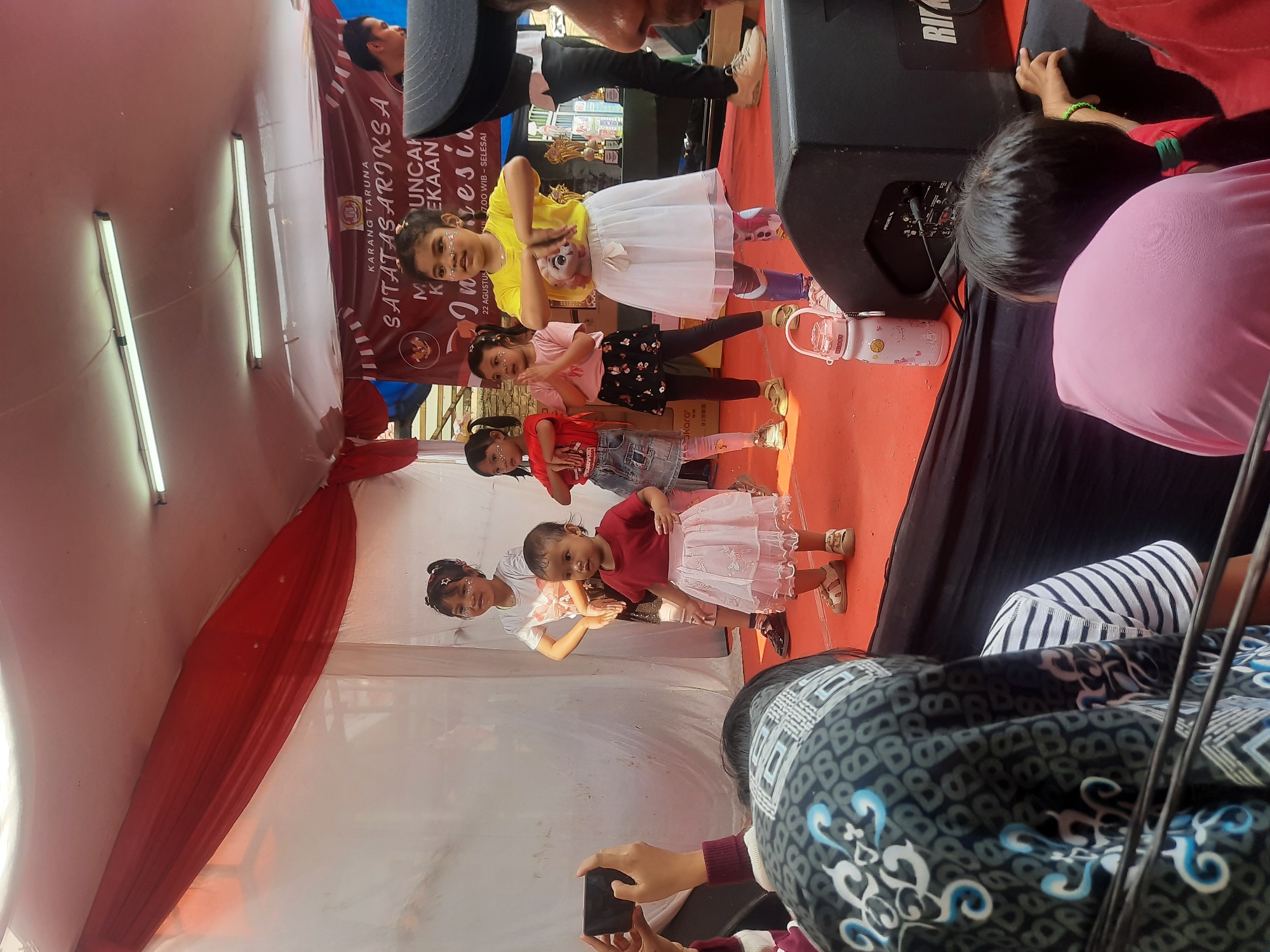 A group of young girls are performing a dance on a stage. They are wearing colorful outfits, including skirts and t-shirts. The stage is decorated with red and white fabric. There are speakers and other equipment on the stage. In the background, a banner with text in Indonesian is visible. Several people are watching the performance from the audience. A person in the foreground is holding a mobile phone, possibly recording the performance. The lighting suggests it is daytime, and the environment appears to be outdoors or in a tent. The banner's text, "KARANG TARUNA SATATASARIKS A MUNCAN KEKAAN Indonesia," suggests a local event, possibly a cultural celebration or talent show, taking place in Bandung City, Indonesia.