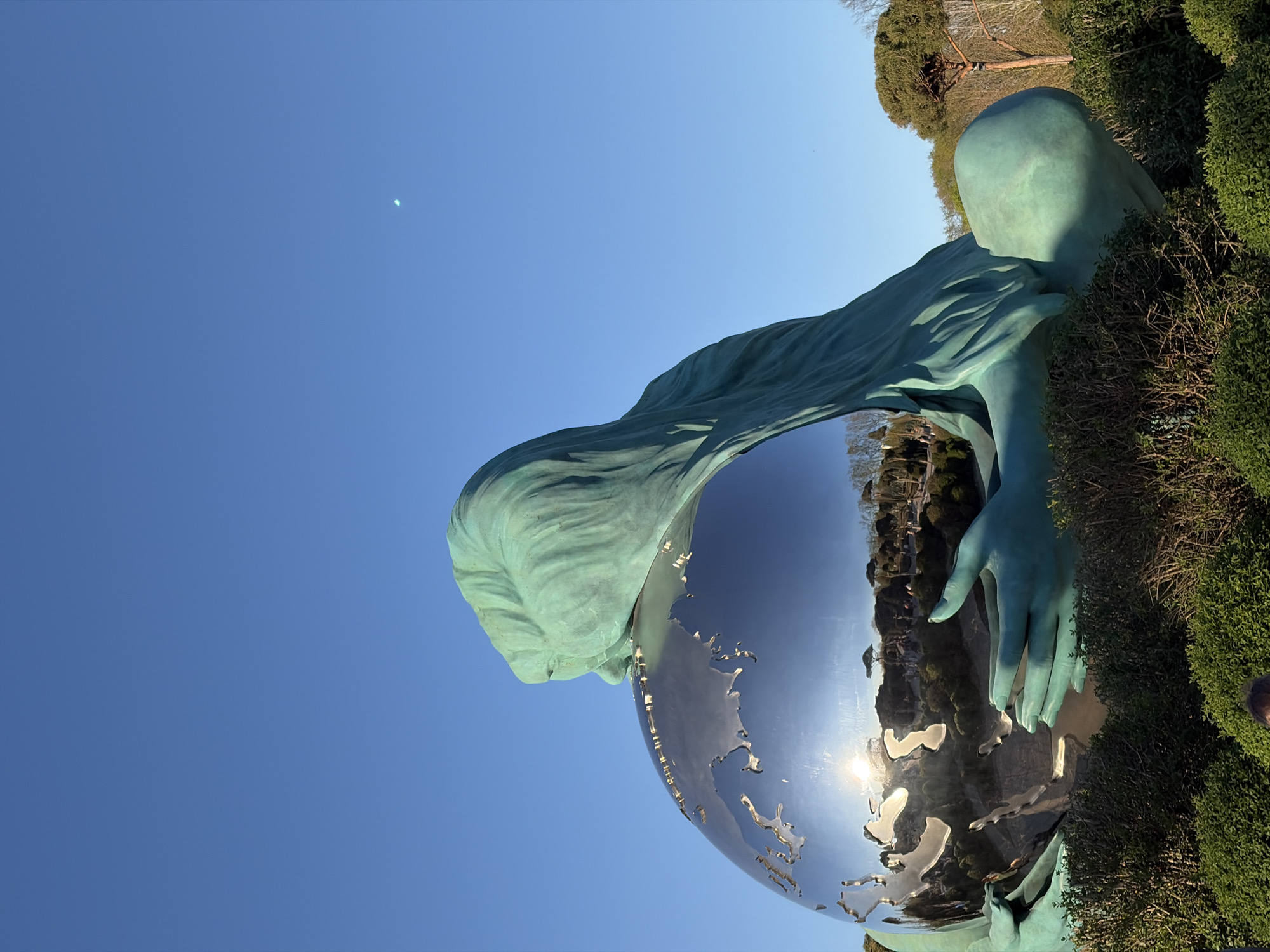 The image features a statue of a woman holding a metallic globe in a park setting. The statue is positioned against a clear blue sky, and the sun reflects off the globe. The woman appears to be made of a blue-green material. Lush green bushes surround the base of the statue, with some trees visible in the background. The globe’s reflective surface shows images of the surrounding landscape, including building and trees. The statue is of the sculpture "Reconciliazione", by Mario Molinari, in Krasnodar, Russia.