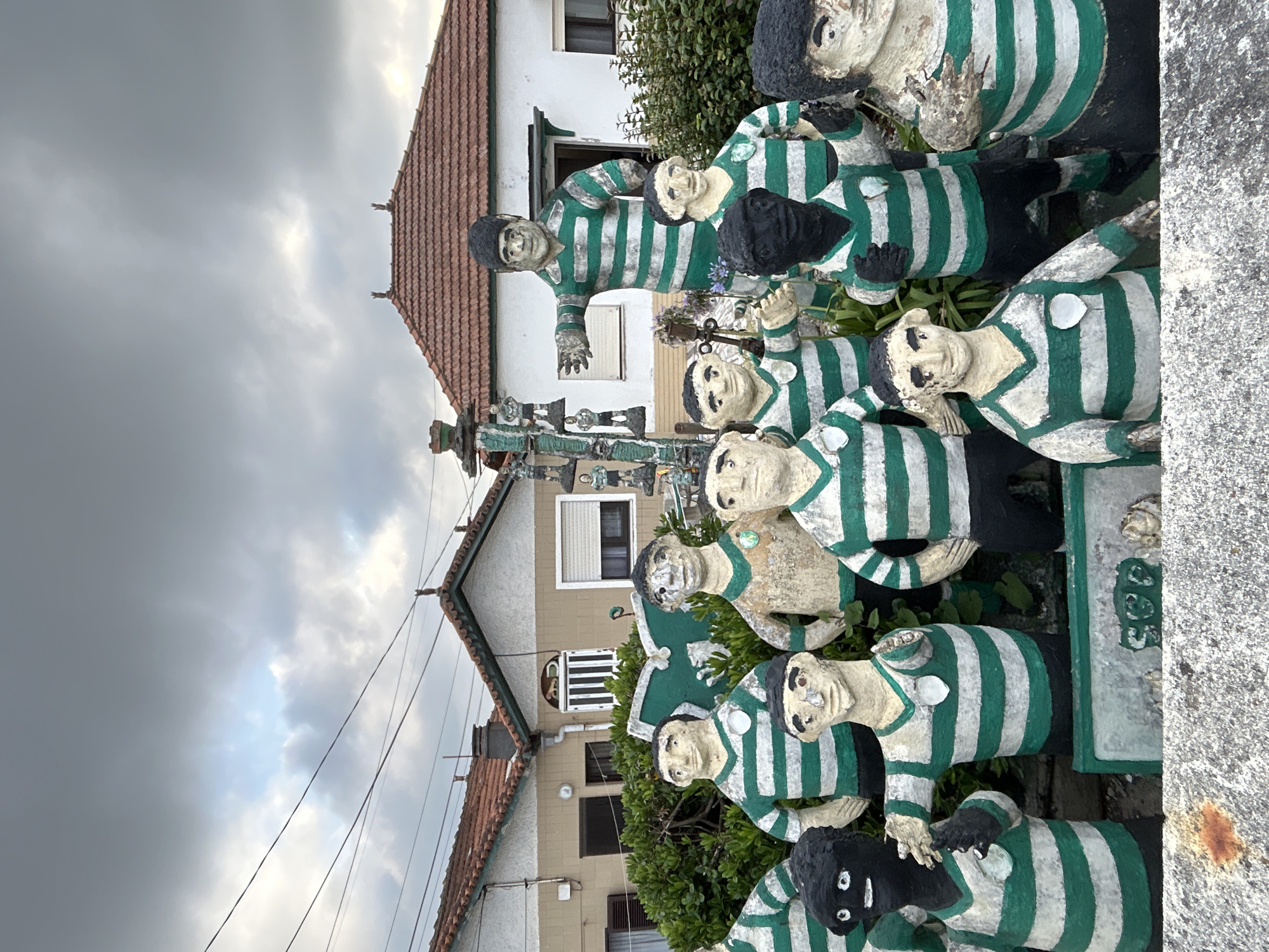 An outdoor, low-angle, medium shot captures a group of statues resembling soccer players adorned in green and white striped jerseys and black shorts. The statues are arranged in clusters, with some standing taller than others, and are positioned in front of a row of buildings with red tiled roofs. The sky above is overcast with dramatic clouds. Some statues have a greenish hue, likely due to weathering. A stone monument, also decorated with green and white elements and featuring smaller statue figures, stands to the left of the main group. The foreground consists of a rough, grey stone surface.
