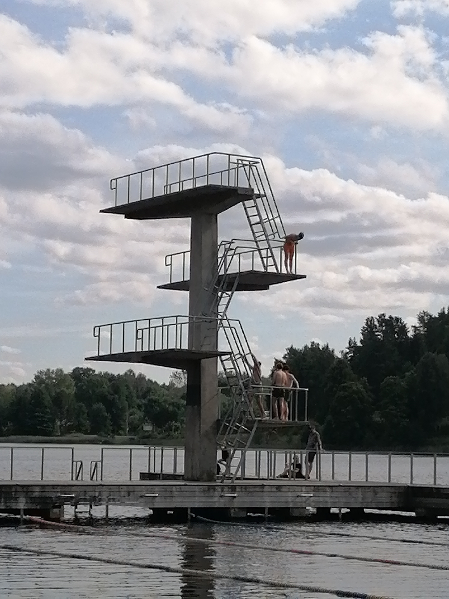 The image shows a diving platform at Lake Zarasas in Zarasai, Lithuania. The platform has multiple levels, with ladders providing access to each. Several people are visible on and around the platform. One person is at the edge of the highest platform, appearing to be preparing to dive. Others are gathered on a lower platform, and some are on a wooden dock extending into the lake. The lake is calm, with visible lane ropes indicating it is used for swimming. The background features a dense line of trees and a cloudy sky.