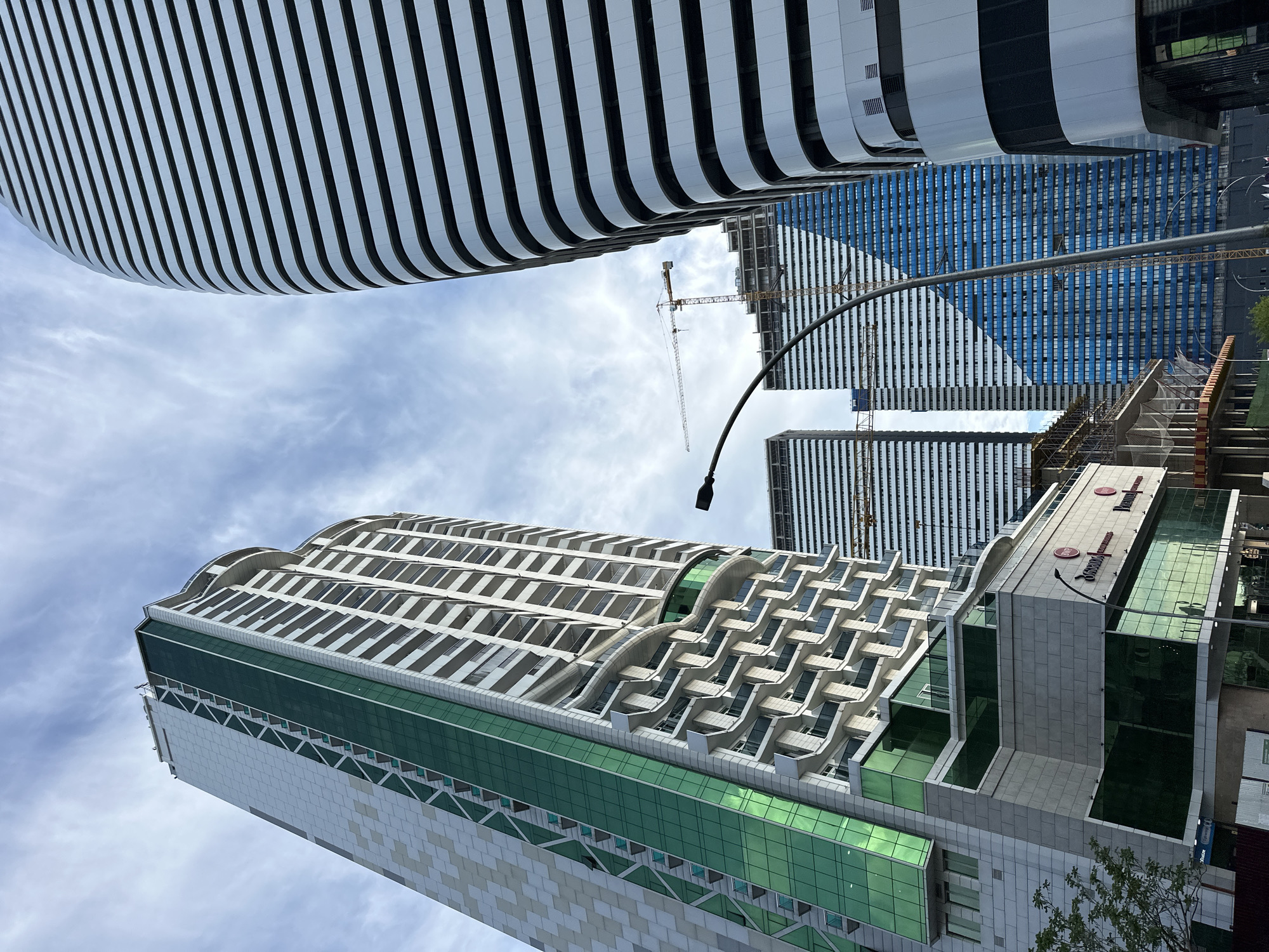 The image captures a low-angle perspective of several high-rise buildings in Batumi, Georgia, against a partly cloudy sky. In the upper-left corner is a curved building with white horizontal lines. Adjacent to this, further in the background, are two taller blue and white buildings with a construction crane visible between them. In the lower-center, a multi-tiered building with green glass panels stands prominently, featuring unique architectural elements on its upper floors. At the bottom of the frame, on the right, is the side of another building with signage in Cyrillic. A street light pole stretches up into the sky, appearing silhouetted against the building facades. The overall scene is one of modern urban development.