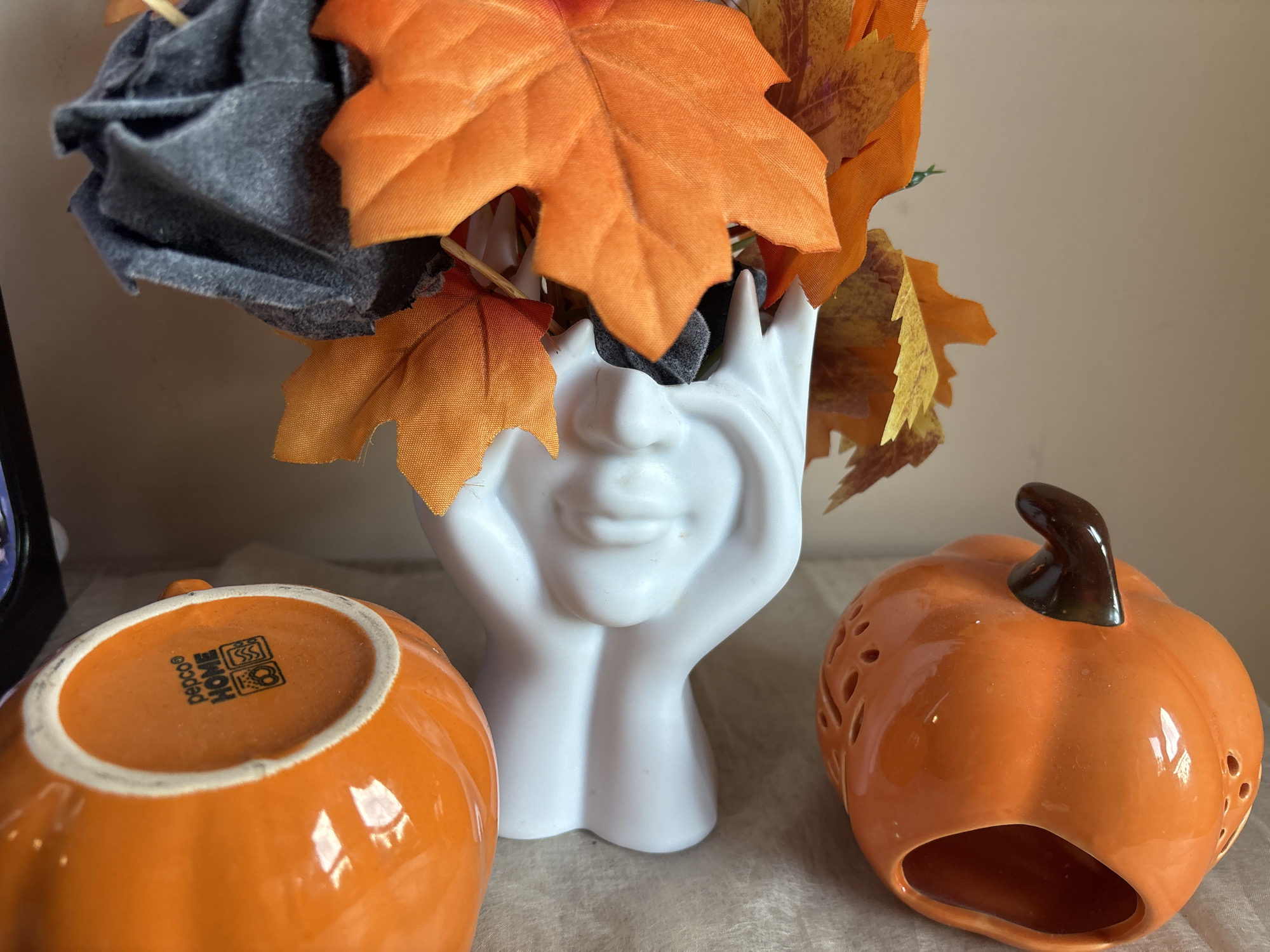 The image shows a close-up of a decorative arrangement. In the center, there is a white ceramic vase shaped like a face with hands placed on the cheeks. The vase is filled with artificial fall foliage, including orange and yellow leaves, as well as some dark gray fabric. To the left of the vase is an upside-down orange ceramic pumpkin with the "pepco HOME" logo visible on its base. To the right of the vase is another orange ceramic pumpkin with carved details and a brown stem. The scene is set on a neutral-colored tablecloth or surface.
The location in the city of Plovdiv, Bulgaria, cannot be determined from this image.