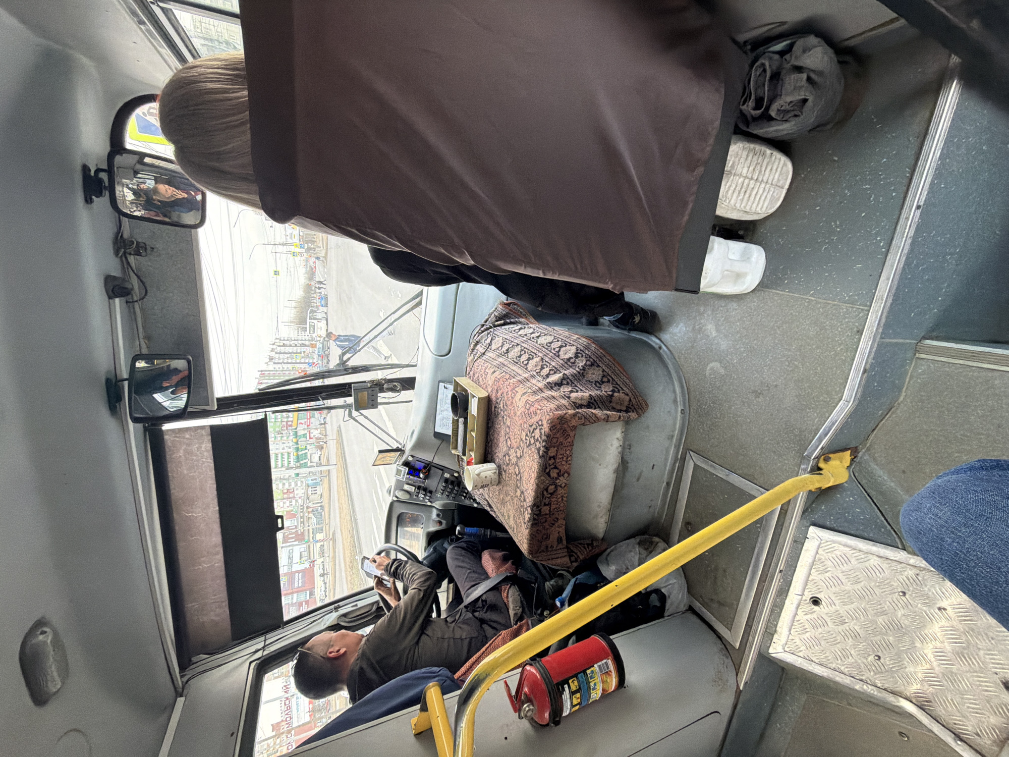The image depicts the interior of a bus in Krasnoyarsk, Russia. The driver, a man wearing glasses and an olive-green shirt, is sitting behind the wheel and looking at a smartphone. A fire extinguisher is located near him. A passenger is sitting behind the driver, partially obscured by a brown blanket or garment. The passenger's feet are visible and wearing different shoes. The dashboard is covered with a patterned textile. A yellow safety bar is nearby. The bus's side window displays a view of buildings. The bus mirror reflects a woman.