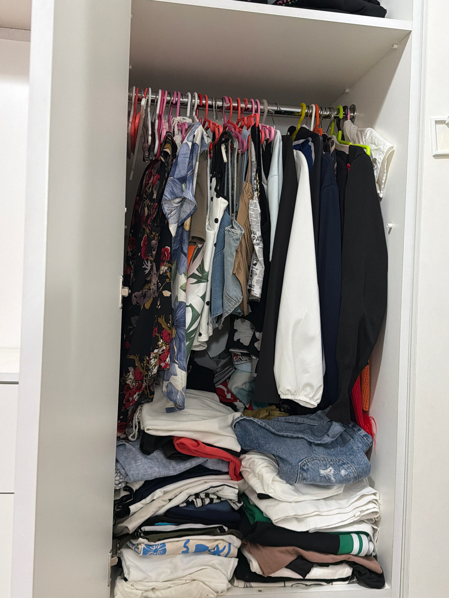 This image shows the interior of a white wardrobe filled with clothes. The upper section of the wardrobe contains clothes hanging on a metal rod, supported by various colored plastic hangers. These hanging clothes include patterned shirts, blouses, and what appear to be dresses. The lower section of the wardrobe is filled with folded clothes stacked on shelves. These folded clothes include t-shirts, denim items, and other garments. The overall impression is of a somewhat disorganized closet. There is no indication of the city of Digah, Azerbaijan or any specific location within it. The image is a close-up view of the wardrobe's contents.