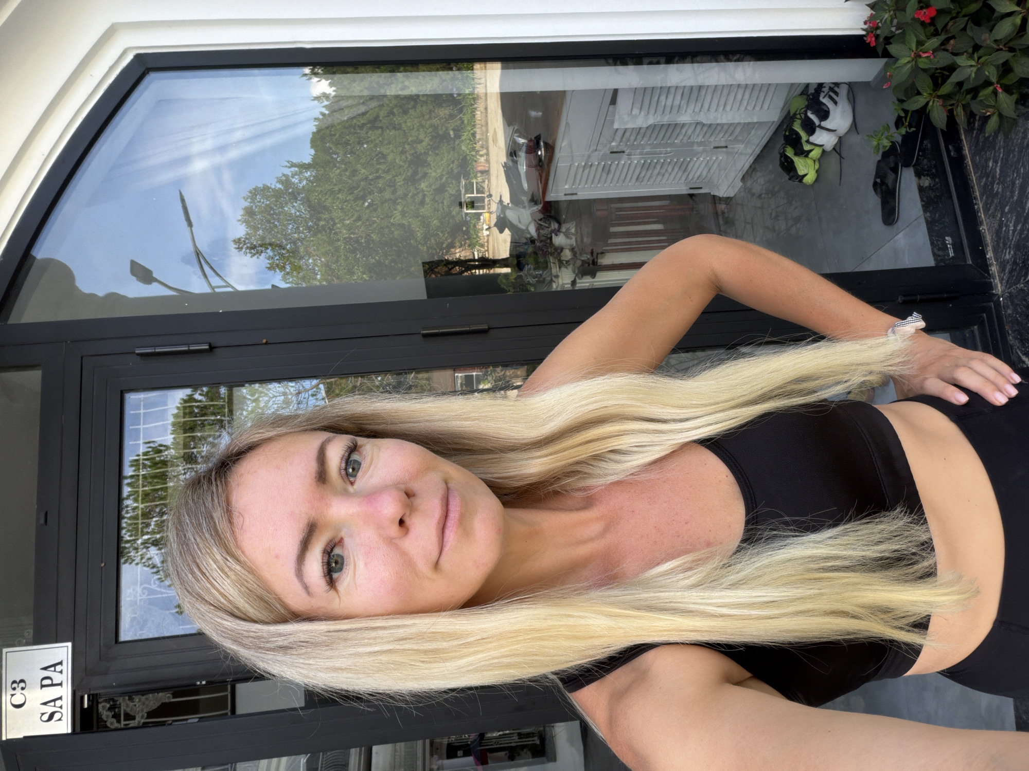 A woman with long blonde hair and fair skin, wearing a black sports bra, stands in front of a large, curved glass window. She is taking a selfie, her left arm extended and her hand out of frame. Her right hand is on her hip. Reflected in the window are trees, a street lamp, and a white cabinet. Outside the window, on the ground, are several pairs of shoes, including neon green and white athletic shoes, and black flip-flops. A sign above the door reads "C3 SA PA". The scene appears to be in Xuân Trường - Đà Lạt, Vietnam.