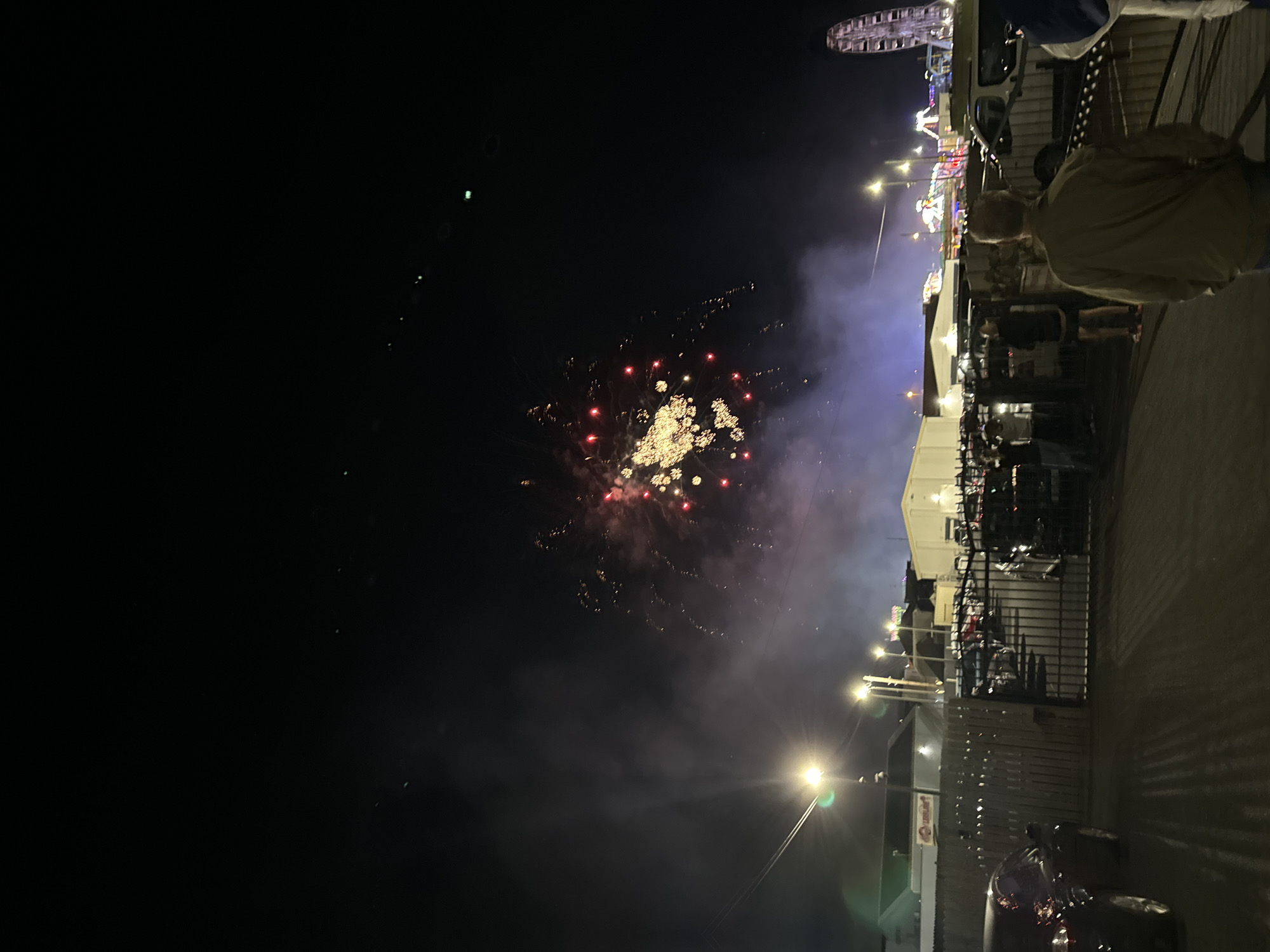 This image depicts a nighttime scene at what appears to be a fairground or outdoor event, possibly the Tramore Amusement Park in Tramore, Ireland, given the presence of a Ferris wheel. Fireworks are actively being displayed in the dark sky, with bright bursts of yellow and red light and trails of smoke. Below the fireworks, the environment is illuminated by streetlights and lights from the amusement park attractions. In the foreground, a crowd of people are visible, some standing and watching the fireworks, while others are walking. There are also cars parked in an area to the left of the frame, and a fence separates the viewing area from the amusement park. The overall atmosphere suggests a festive and celebratory event.