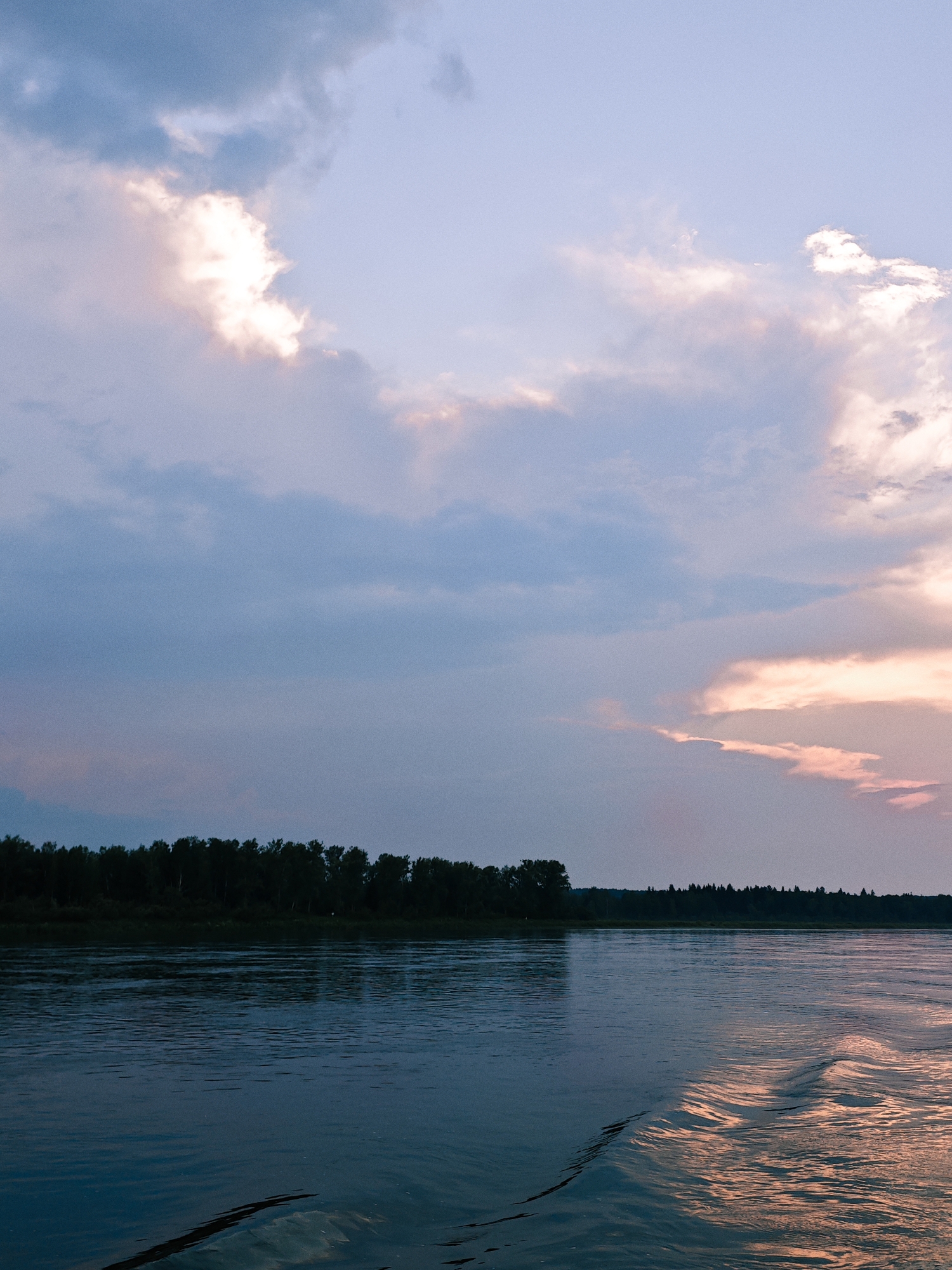 This is a photograph taken from a body of water, likely a river, looking towards a tree-lined shore under a twilight sky. The sky is filled with clouds, some of which are illuminated with soft pink and white hues, indicating the setting or rising sun. The water is dark and reflects the sky, with visible ripples and a wake trailing from the perspective of the viewer, suggesting movement. The shore is densely forested with dark green trees, forming a natural horizon. The scene is serene and atmospheric, with no people or man-made structures immediately visible. The location is identified as Kazachinskoe, Russia.