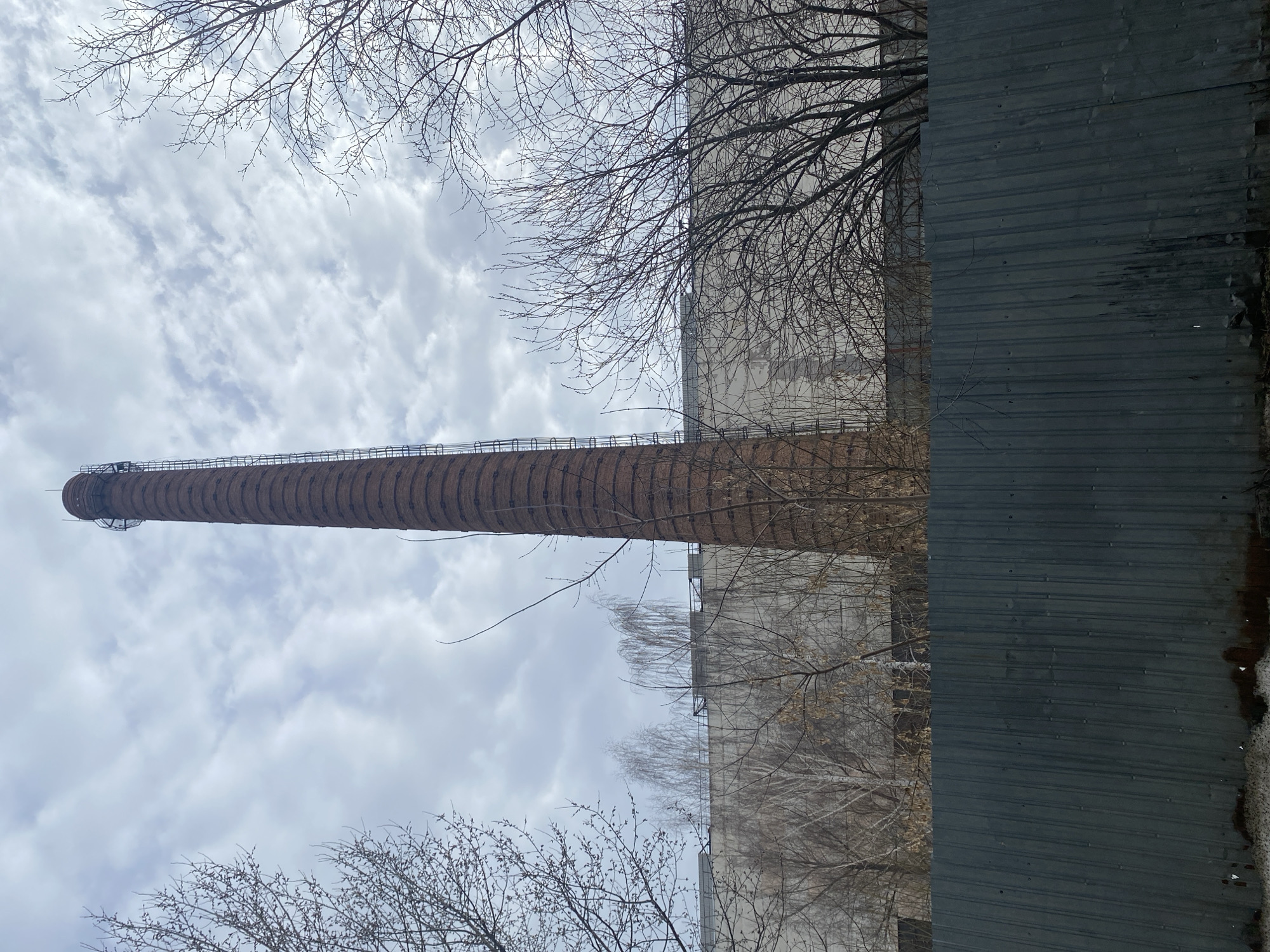An outdoor, tilted long shot captures a tall brick smokestack, presumably industrial, extending into a sky filled with white and gray clouds. Bare branches of trees are visible in the foreground and partially obscure the view towards the upper right and upper left of the frame. A large, concrete building is situated to the right of the smokestack, and a grey corrugated metal wall runs vertically down the right edge of the frame. The location is the city of Izhevsk, Russia.