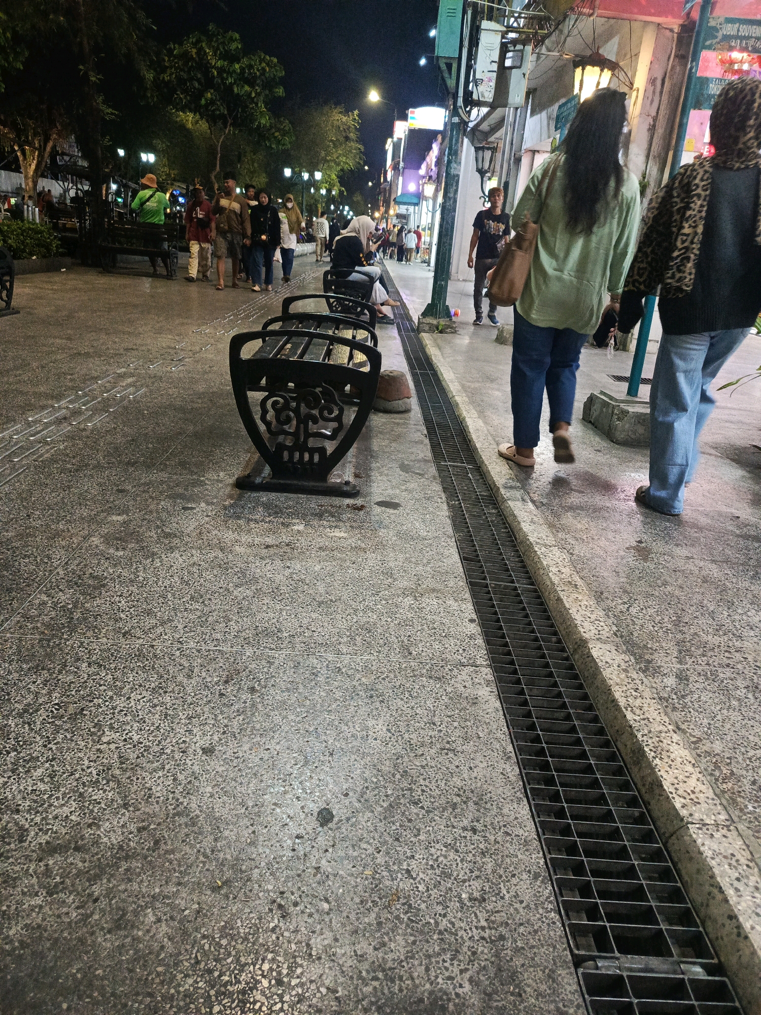 This is a nighttime, eye-level, medium shot of Malioboro Street in Yogyakarta, Indonesia. The street is paved with textured concrete, with raised tactile paving lines indicating a pedestrian walkway. A metal grate drain runs along the right side of the walkway. In the foreground, a black metal bench with an ornate design is visible.
Several people are walking away from the camera on the walkway. Two individuals are most prominent: one on the left wearing a light green long-sleeved shirt and blue jeans, and another on the right wearing a black jacket with a leopard print pattern and blue jeans. A third person, a male wearing a dark t-shirt and pants, is walking towards the camera in the background. Further back, a group of people are walking, and one person in a green shirt and a straw hat is seated on a bench. Trees and lampposts line the street, with illuminated shop signs visible in the background. The overall scene depicts a busy urban street at night.