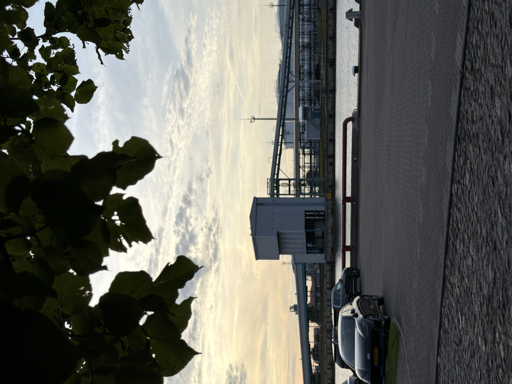 The image captures a view of the Ventspils Port industrial area in Ventspils, Latvia. In the foreground, two cars are parked on a cobblestone and paved surface. The car on the left is a dark sedan, and behind it is a dark SUV. A body of water is visible beyond the parking area, separated by a red railing. On the opposite side of the water, a large industrial building with a grey structure and conveyor belts is prominent. In the background, more industrial facilities and storage tanks are visible against a cloudy sky with warm sunset light. Silhouetted tree leaves frame the top portion of the image.