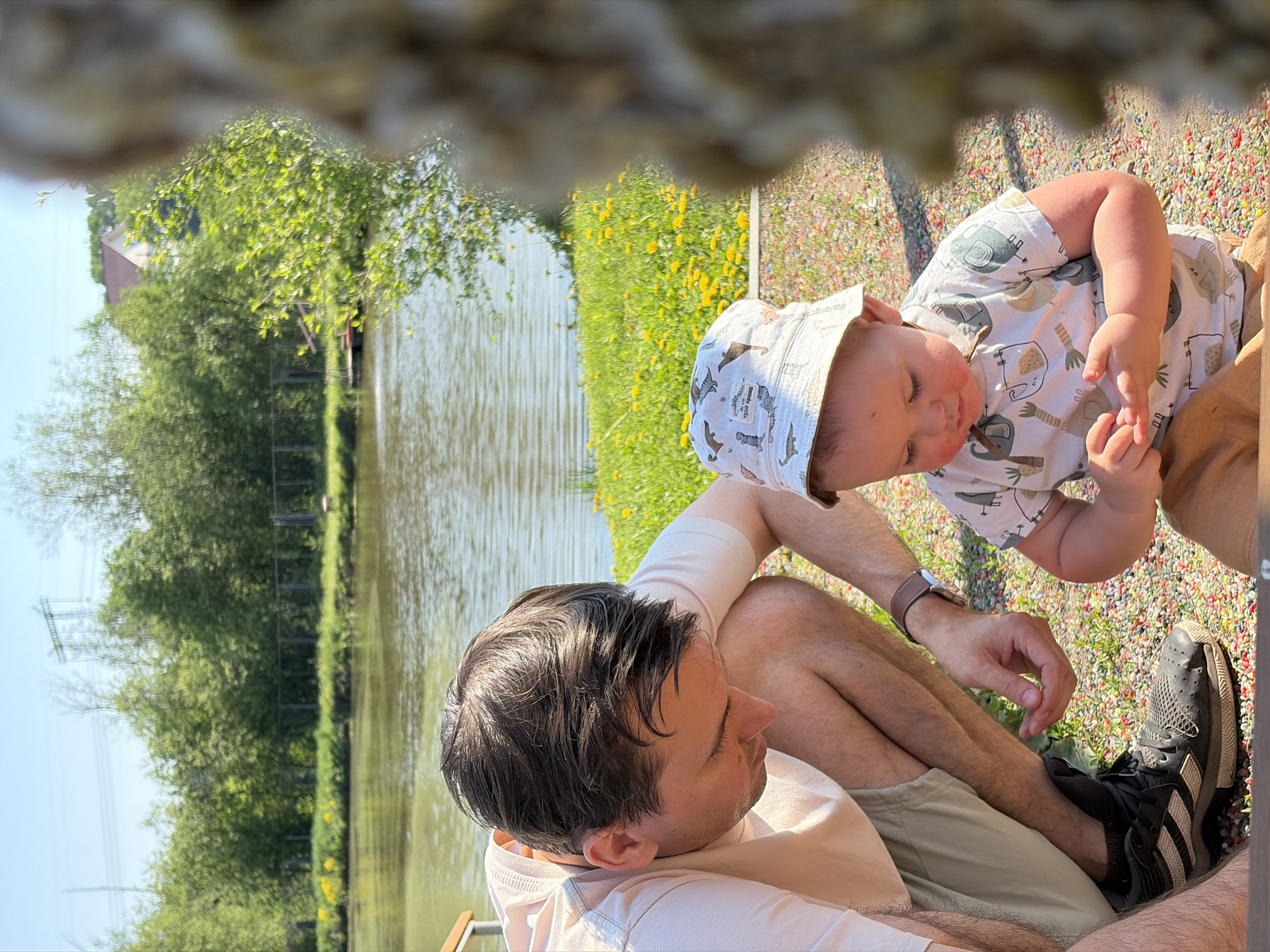 The image shows a man and a baby outdoors in Malyi Bukor, Russia, next to a body of water. The man is sitting on the ground with his legs crossed, wearing a light-colored t-shirt, tan shorts, and dark-colored sneakers. He is looking at the baby. The baby is wearing a patterned hat and a short-sleeved shirt with elephant patterns, and is touching the man's hand. The ground is covered in colorful pebbles and some grass. In the background, there is a body of water with green vegetation along the shoreline. Power lines and a building can be seen in the distance. The scene is shot from a high angle, partially obstructed by an out-of-focus object in the upper part of the frame.