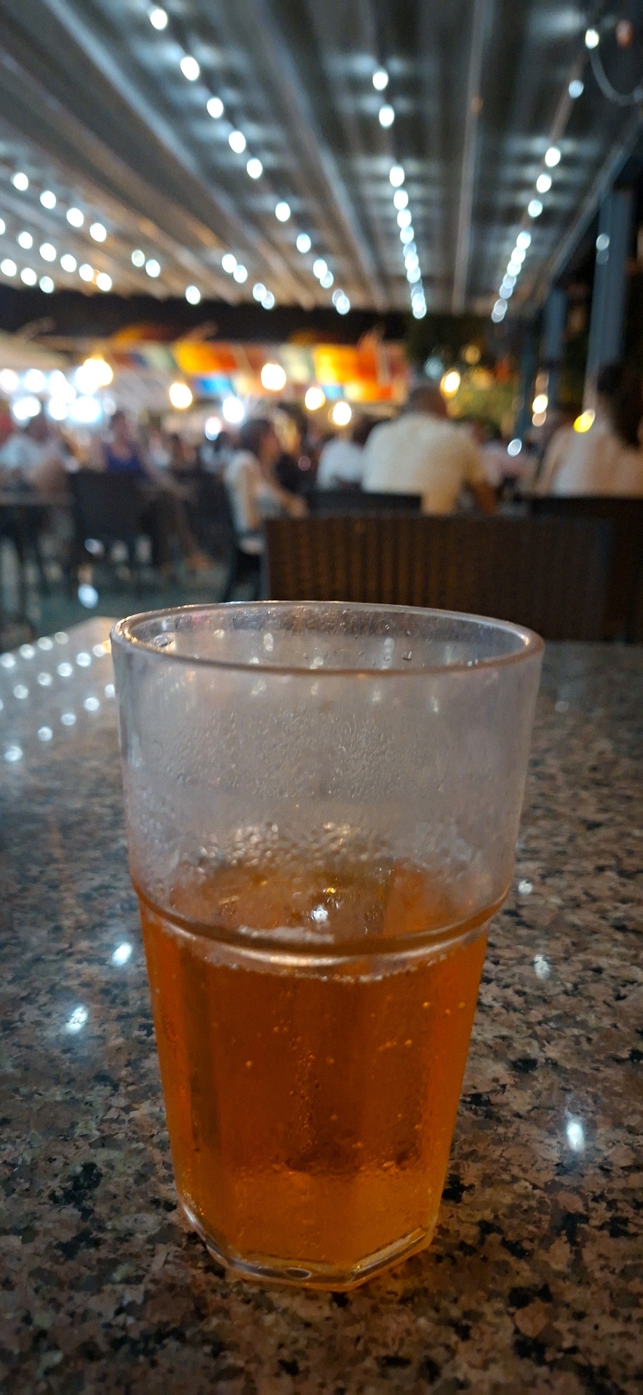 A close-up, low-angle vertical shot captures a glass of amber-colored liquid, likely a beverage, set on a speckled granite tabletop. The glass, partially filled and showing condensation on its exterior, is positioned in the foreground, dominating the frame. Behind the glass, a blurred background depicts an outdoor seating area at night, possibly a restaurant or bar. Strings of small, bright lights are visible, suspended overhead, creating a festive and illuminated atmosphere. Several out-of-focus figures are seated at tables in the distance, suggesting a social gathering. The overall scene conveys a relaxed, evening ambiance. The photograph was taken in Kemer, Turkey.