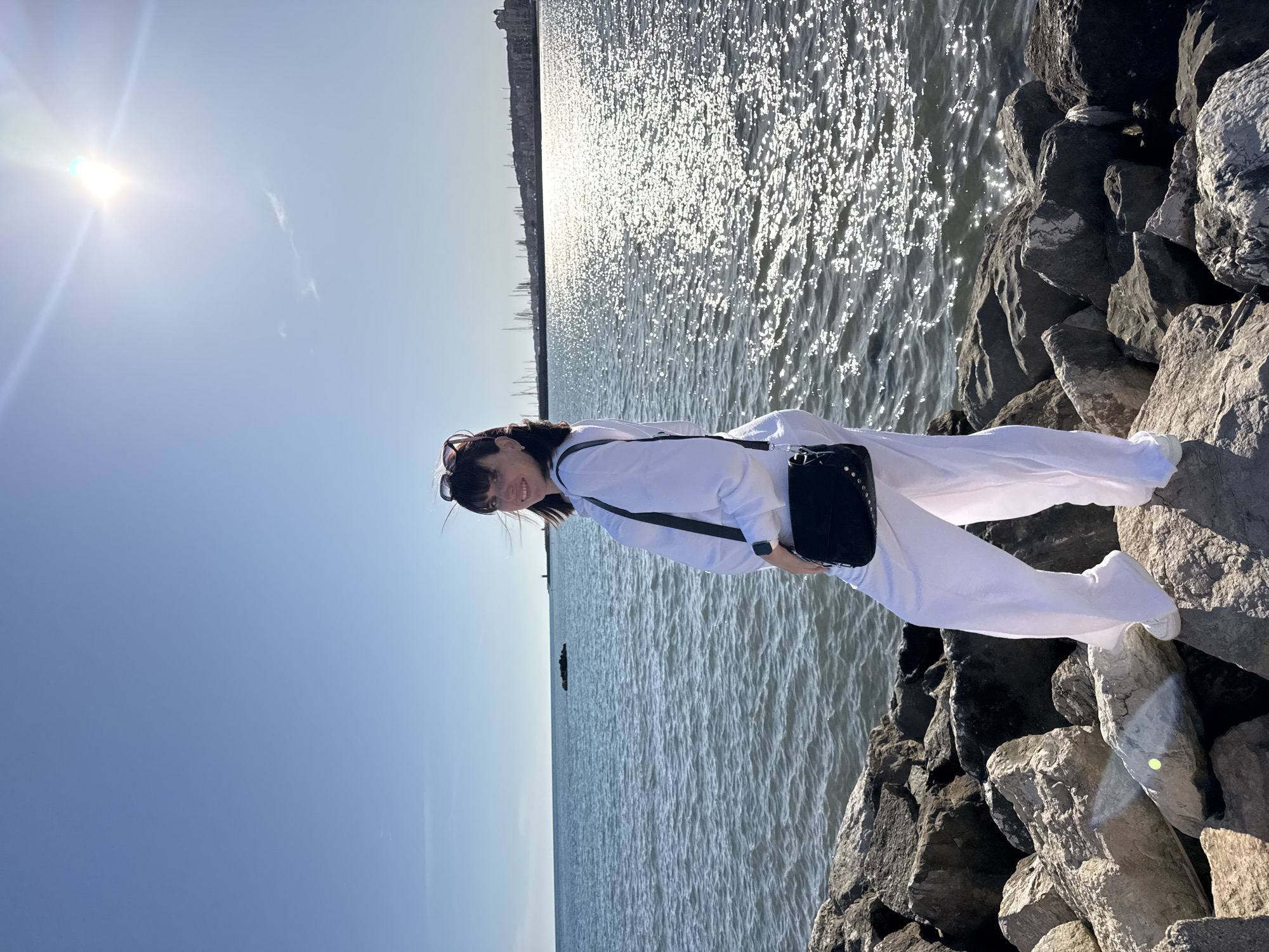 This bright, sunny image, captured in Nettuno, Italy, features a woman posing on large rocks by the sea. The woman, positioned on the right side of the frame, smiles warmly at the camera. She is dressed in a crisp white long-sleeved top and matching white wide-leg pants. A black shoulder bag, adorned with silver studs, hangs across her body, and sunglasses rest on her head.
The scene is dominated by a vast expanse of blue water, which stretches towards a distant horizon. The sun, high in the upper left of the frame, casts brilliant reflections across the water's surface, creating a sparkling effect and indicating a clear, bright day. A faint silhouette of buildings or structures can be seen along the far coastline.
The foreground and midground are composed of large, rugged rocks, likely forming a breakwater or rocky shore, on which the woman stands. The sky above is a clear, light blue, with a noticeable sun flare in the upper left, highlighting the intense sunlight. The overall atmosphere of the image is cheerful and relaxed, reflecting the woman's pleasant expression and the idyllic coastal setting. There is no visible text in the image.