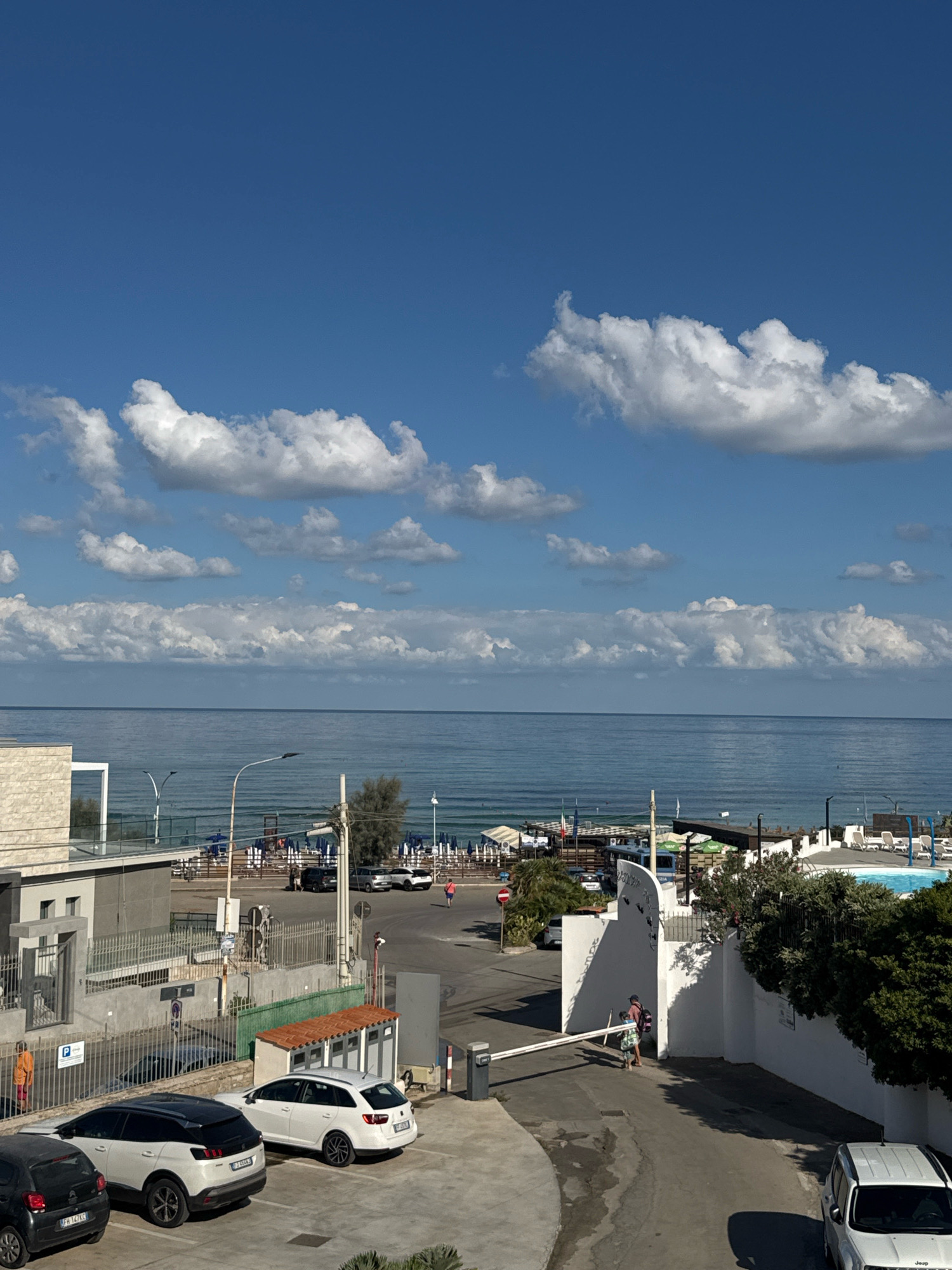 This is an eye-level, medium shot taken on a sunny day, looking towards the sea from a residential or hotel area in Capaci, Italy. The sky is a bright blue, dotted with fluffy white clouds. The sea is calm and blue, stretching to the horizon.
In the foreground, there is a paved area with several parked cars. From left to right, there is a dark grey compact car, a white SUV, and a white station wagon. To the right, a white Jeep SUV is parked facing away from the camera. A white automatic gate is extended across the road in the middle ground.
Beyond the parking area, there is a pathway leading towards the beach. On the left, a light-colored building with a textured facade is visible. There are several smaller structures with orange tiled roofs, possibly restrooms or utility buildings. A fence separates the parking area from the pathway.
Further in the distance, the beach is lined with rows of lounge chairs and umbrellas, indicating a resort or public beach area. A swimming pool with blue water is visible on the far right. A few people are visible walking or standing in the area.
In the middle ground, a few trees are scattered, and there are streetlights and poles. A person in an orange jacket is visible walking near the parked cars on the left. Two people, one in a blue shirt and the other in a checkered shirt, are walking towards the extended gate on the right.