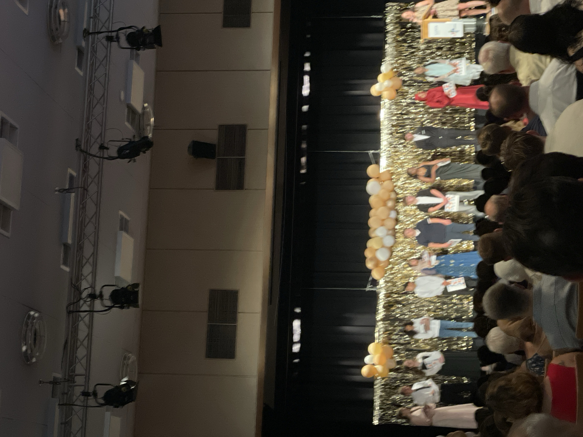 The image depicts an awards ceremony taking place on a stage in what appears to be an auditorium or event hall. Several individuals, likely award recipients, are standing on the stage, posed for photographs. They are dressed in formal attire, with some holding certificates or awards. The stage is decorated with balloons and a shimmering backdrop. In the foreground, an audience is seated, observing the ceremony. The overall atmosphere suggests a celebratory event, possibly a school or organizational awards night. The location is identified as Nidda, Germany.
