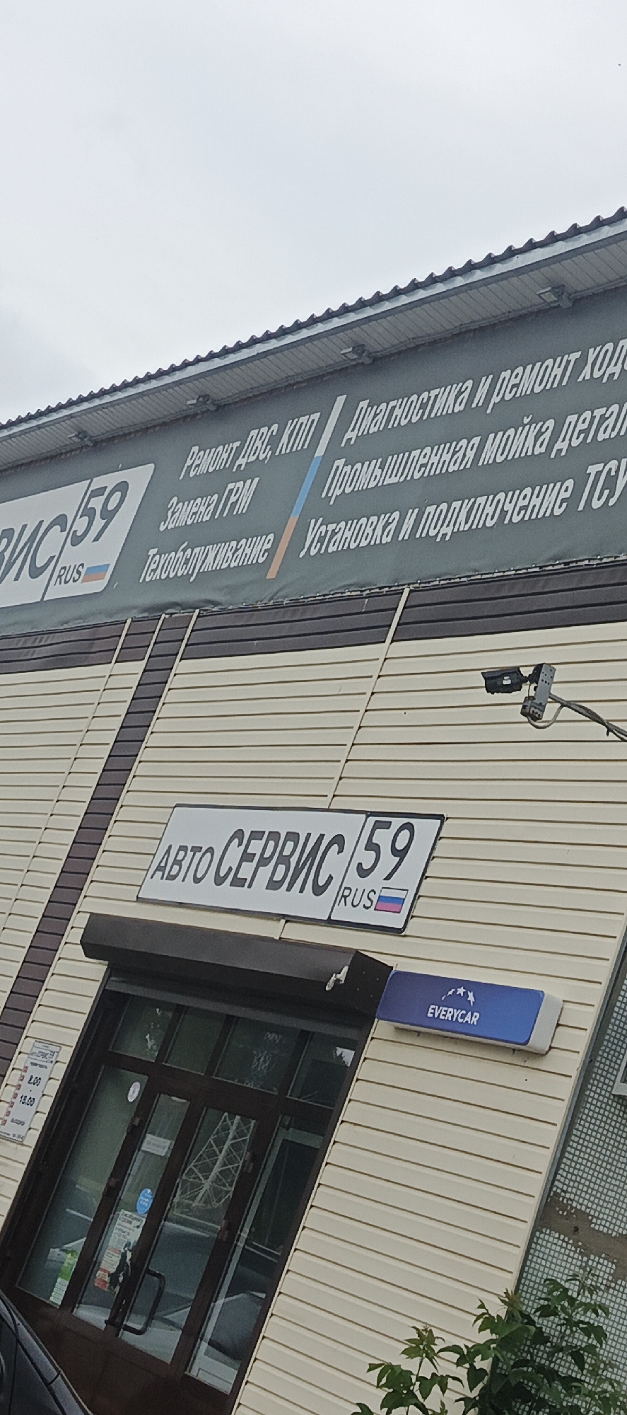 The image shows the facade of a building with a sign that reads "АВТО СЕРВИС 59" and "RUS" with the Russian flag. The building is clad in beige siding with dark brown trim. A security camera is mounted on the wall above the sign. Below the "АВТО СЕРВИС 59" sign, there is another sign that says "EVERYCAR" in white text on a blue background with three white stars. The entrance to the building is a glass door with dark brown framing, and posters are visible through the glass. To the left of the door, there is a sign with Russian text and a clock displaying times from 8.00 to 18.00. The overall impression is that of a car service center. The location is identified as Chaikovsky, Russia, due to the "59" in the sign, which likely refers to the Perm Krai region, where Chaikovsky is located.