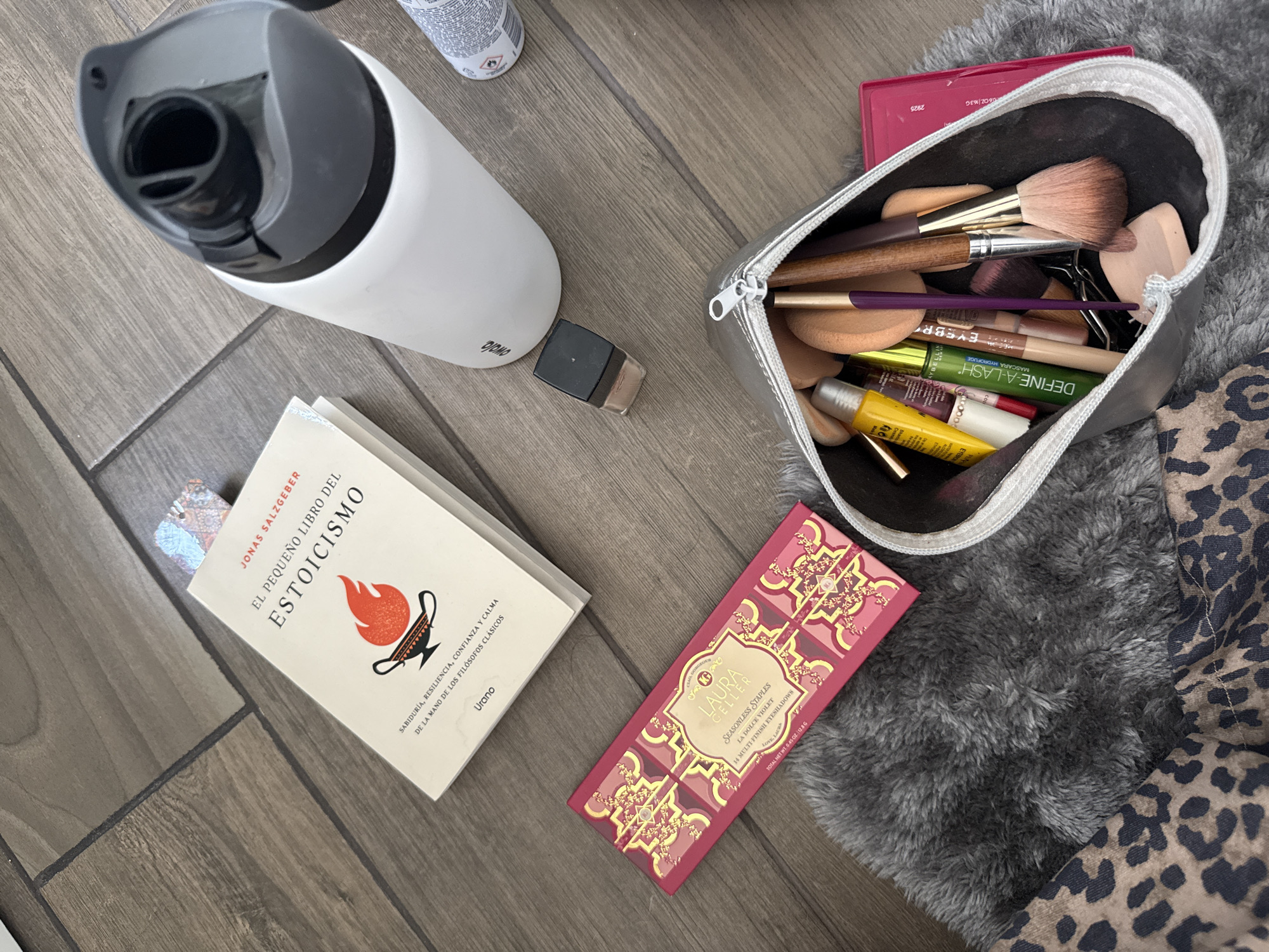 The scene presents a variety of personal items arranged on a wooden surface that resembles a plank floor. A white water bottle with a black lid sits prominently. Next to it, there is a book titled "El Pequeño Libro del Estoicismo" ("The Little Book of Stoicism") by Jonas Salzgeber, with a bookmark sticking out. A small container of makeup is positioned near the bottle.
A silver makeup bag, filled with an assortment of makeup brushes, sponges, and tubes of products, is adjacent to a Laura Geller eyeshadow palette in shades of pink and gold. The makeup bag rests partially on a gray, fluffy rug, which is itself next to a garment with a leopard print.