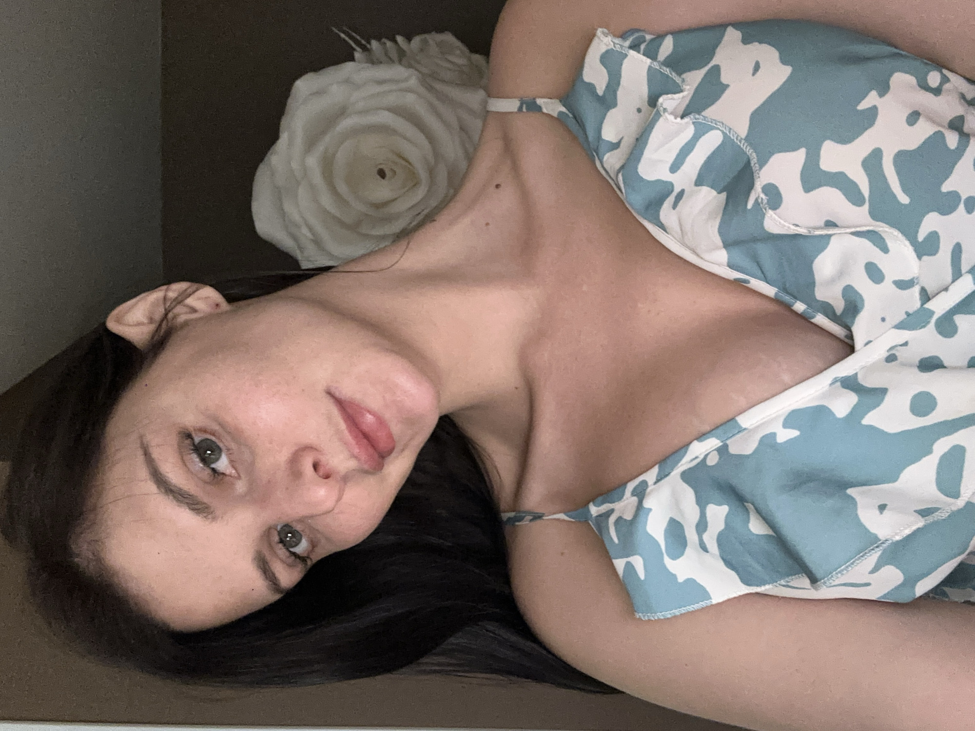 A young woman with long, dark hair and green eyes looks directly at the camera, her head tilted slightly. She is wearing a blue and white patterned dress with ruffled straps. Her skin appears fair with a scattering of freckles. Behind her, a large, white rose decoration is visible. The background is dark and out of focus. The image is a close-up portrait, likely taken indoors.