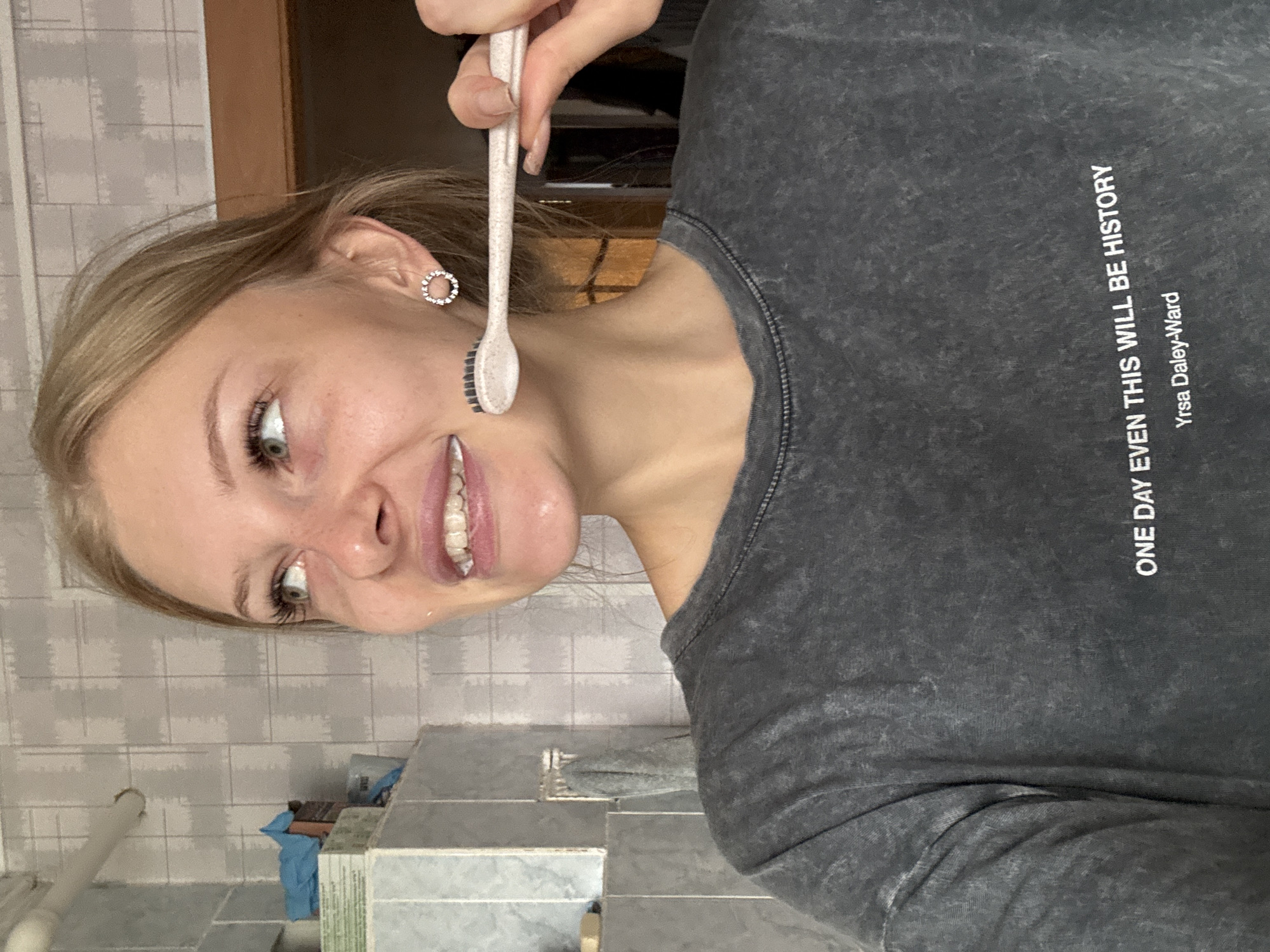 A young woman with blonde hair and light-colored eyes is in a bathroom, brushing her teeth with a pink toothbrush. She is wearing a dark gray t-shirt with white text that says "ONE DAY EVEN THIS WILL BE HISTORY" and "Yrsa Daley-Ward." The bathroom has tiled walls. The scene appears to be in a residential bathroom. No specific location in Borodulino, Russia is identifiable from the image.