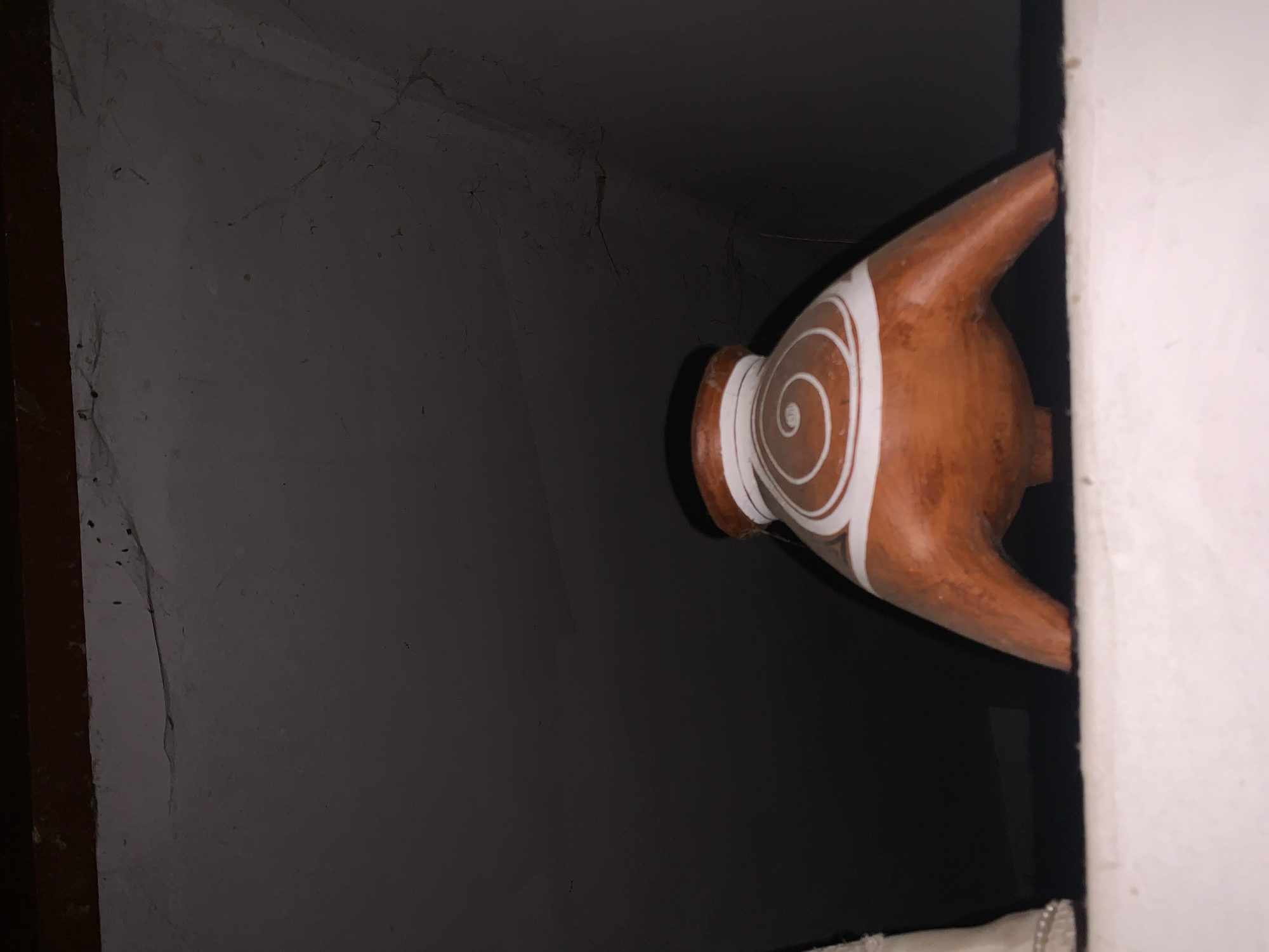 An indoor view presents an orange-brown, vase-like object featuring white spiral decoration, resting close to a white wall. The vase is positioned in a dim or poorly lit space, possibly within an alcove or niche, given the dark wall behind it. The image does not provide any specific location details or further details about the city.